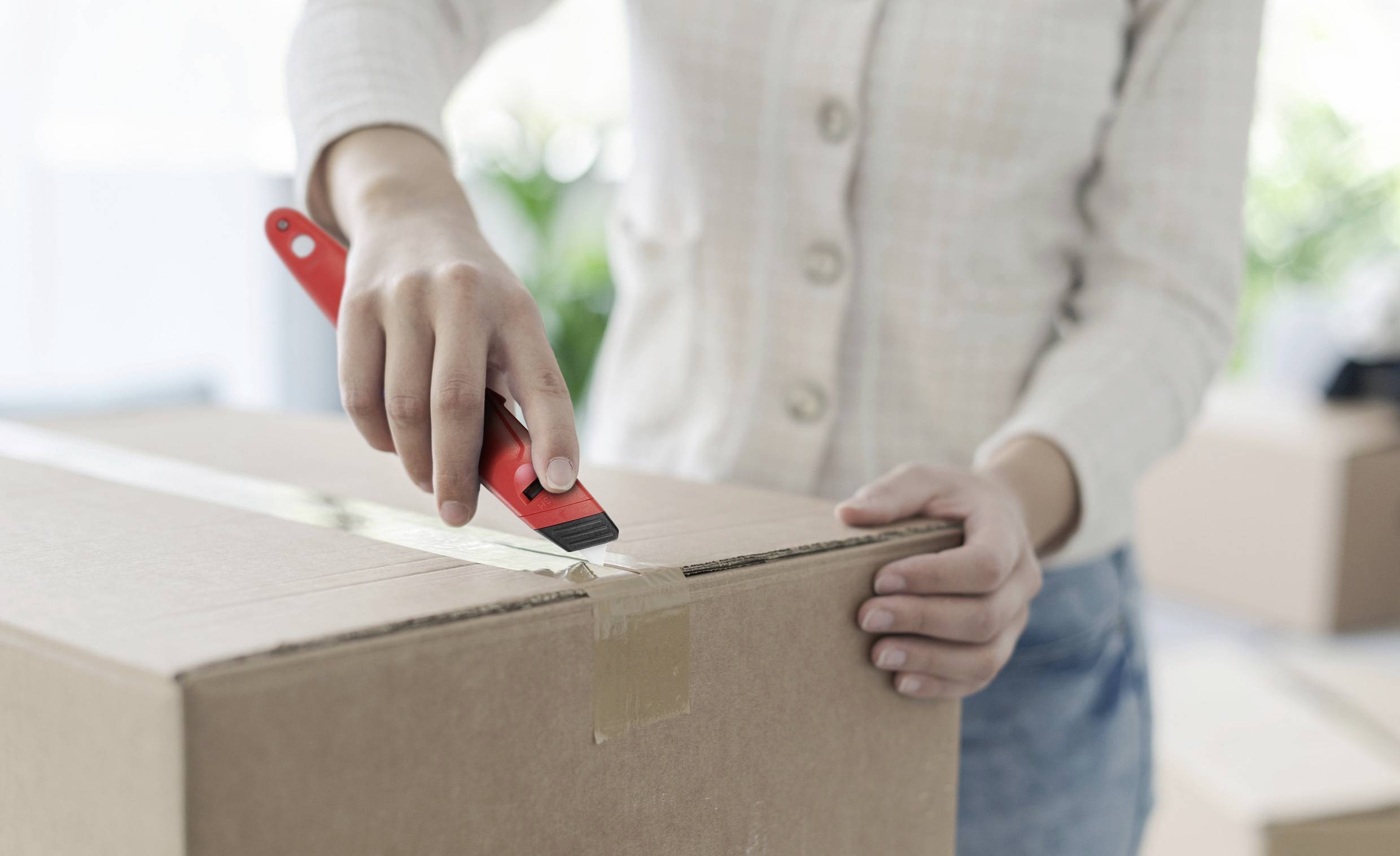 Une personne ouvre un grand carton avec un cutter à lame rouge. L'arrière-plan montre des boîtes emballées de manière floue.