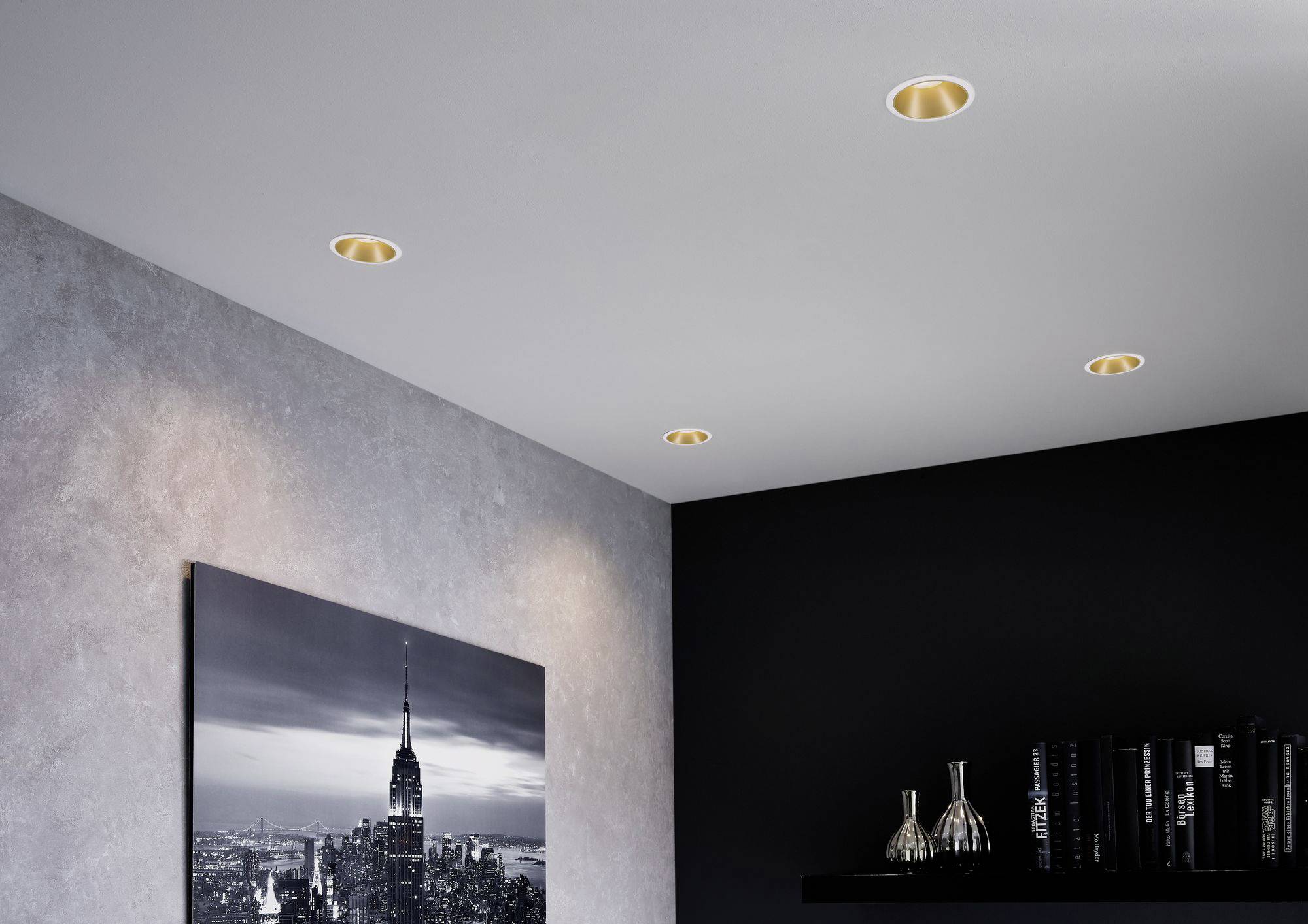Quatre spots encastrés au plafond éclairent une chambre à coucher. Un photographie noir et blanc d'une ville est suspendue au mur. Une étagère avec des objets décoratifs est visible.