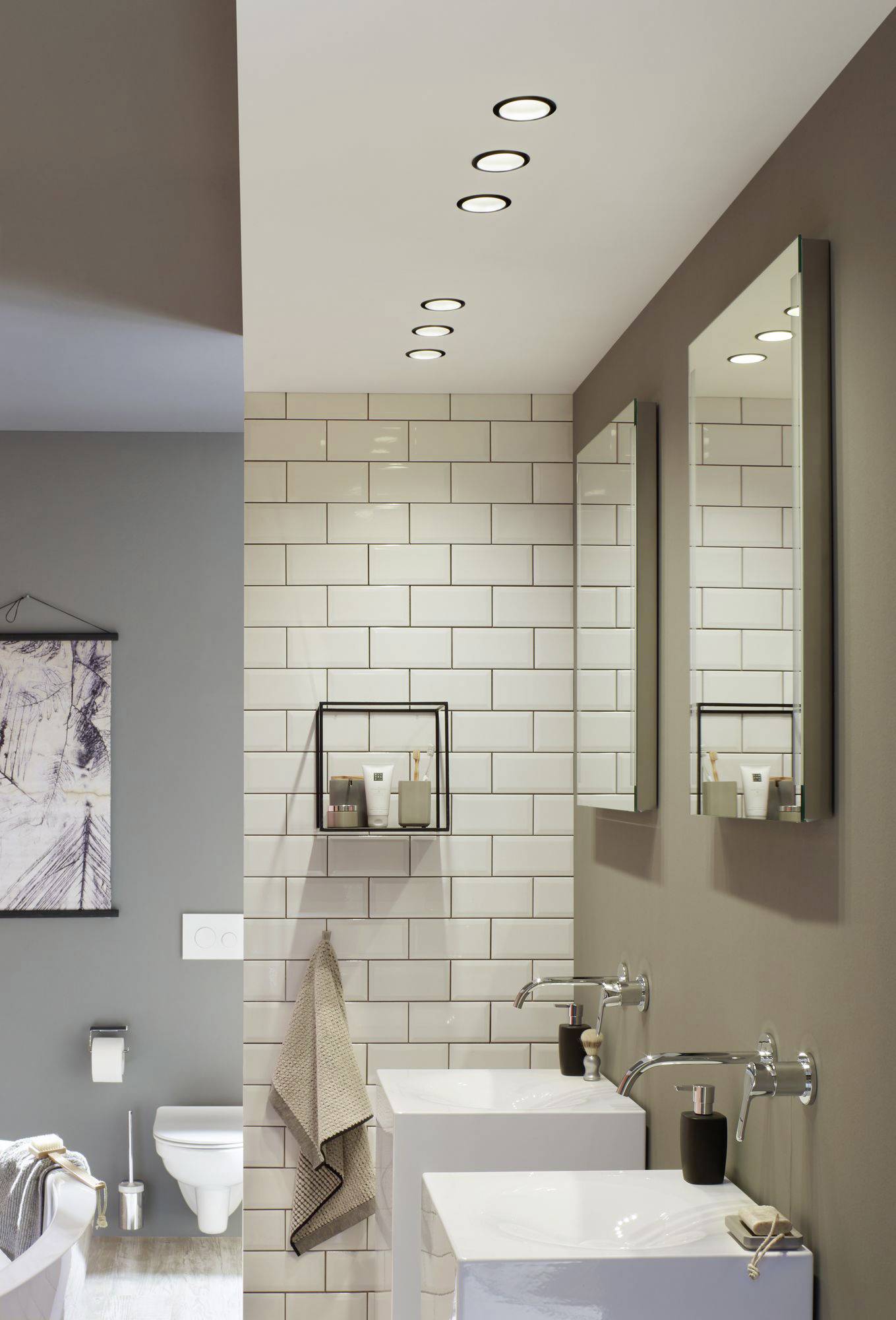 Salle de bain moderne avec carrelage blanc, deux lavabos avec miroirs, étagère murale avec produits de soin et vue arrière sur les toilettes.