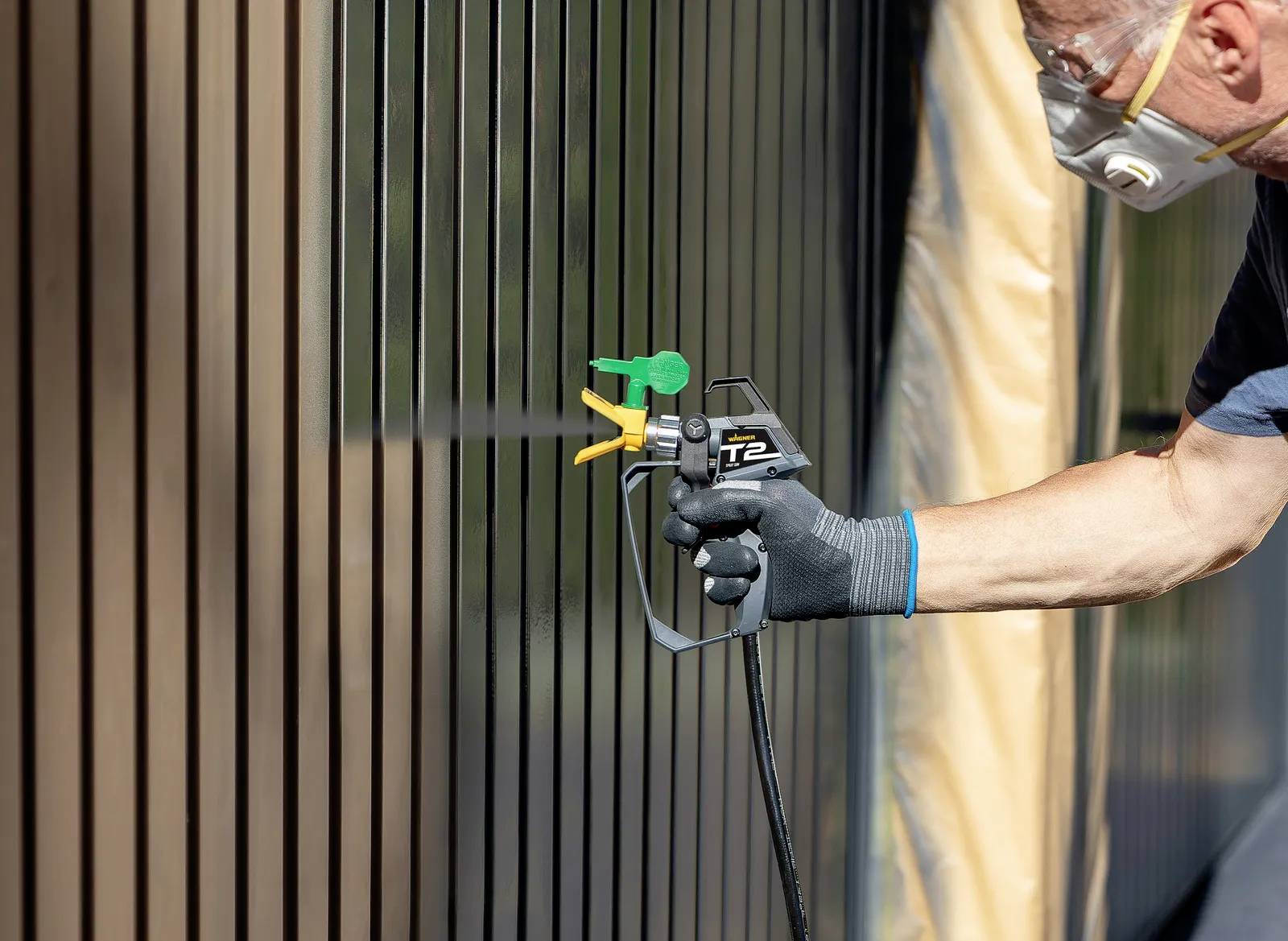 Une personne pulvérise de la peinture sur un mur métallique à l'aide d'un pistolet de peinture. Elle porte un masque de protection et des gants.