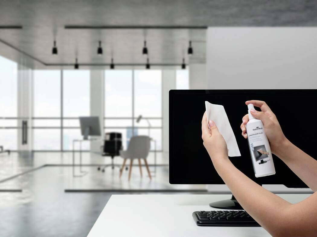 « Une main tenant un vaporisateur étiqueté « Nettoyant pour écran » et un chiffon nettoie un écran d'ordinateur. Un espace de bureau moderne est visible en arrière-plan. »