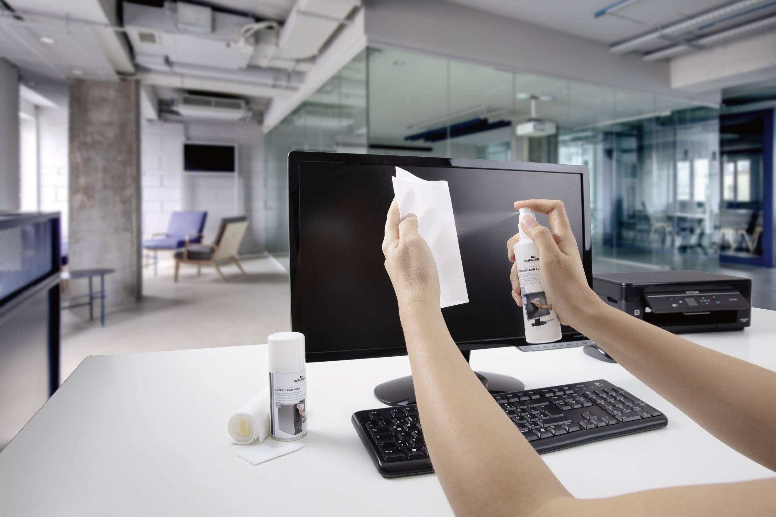 Une personne nettoie un écran d'ordinateur avec un chiffon et un spray de nettoyage dans un bureau moderne avec plusieurs postes de travail.