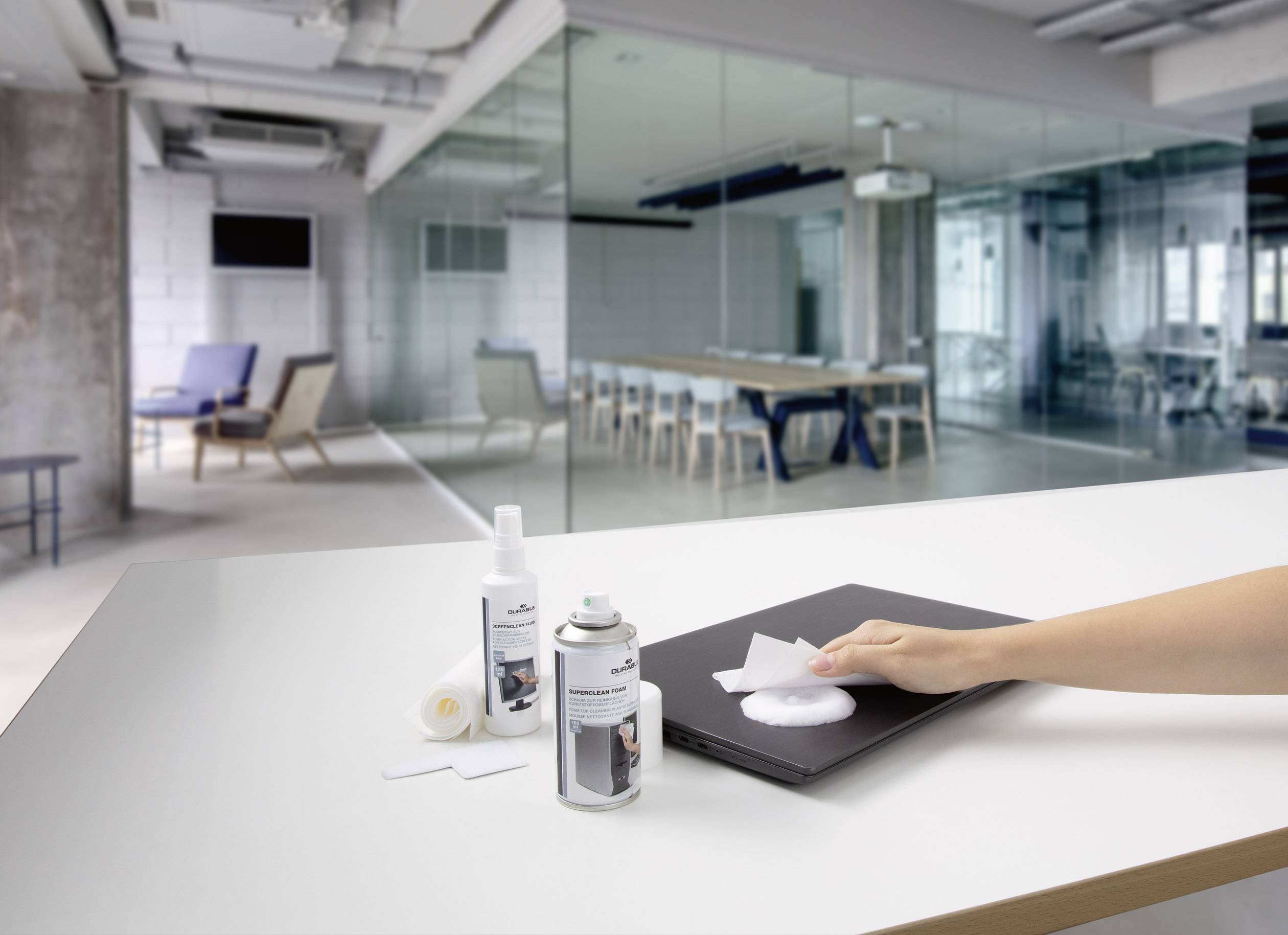 Une main nettoie un ordinateur portable avec un chiffon et un spray désinfectant sur un bureau dans un bureau moderne, avec une salle de réunion en arrière-plan.