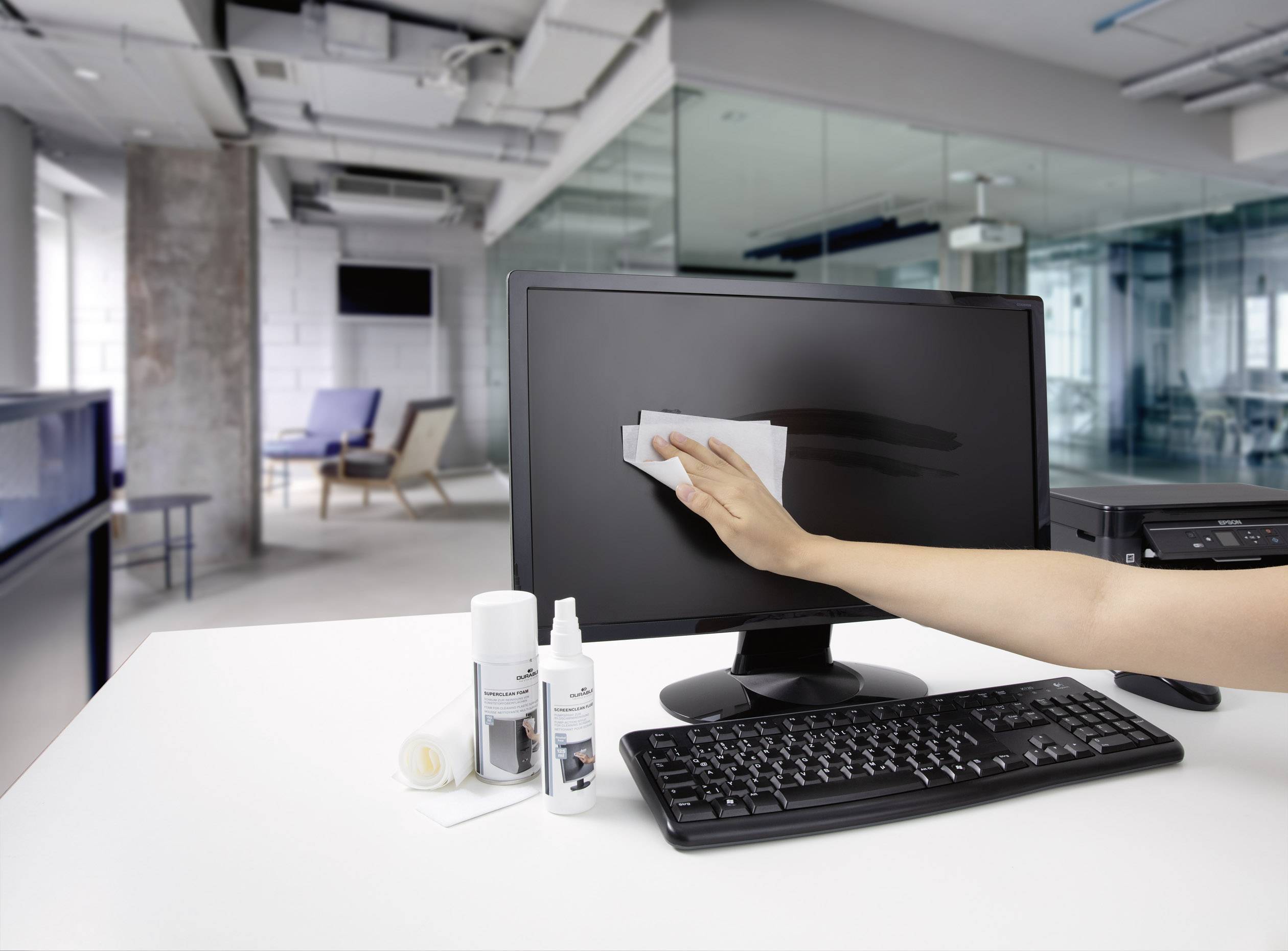 Une main nettoie l'écran d'un ordinateur avec un chiffon dans un bureau moderne. Table avec clavier, moniteur, produits de nettoyage.