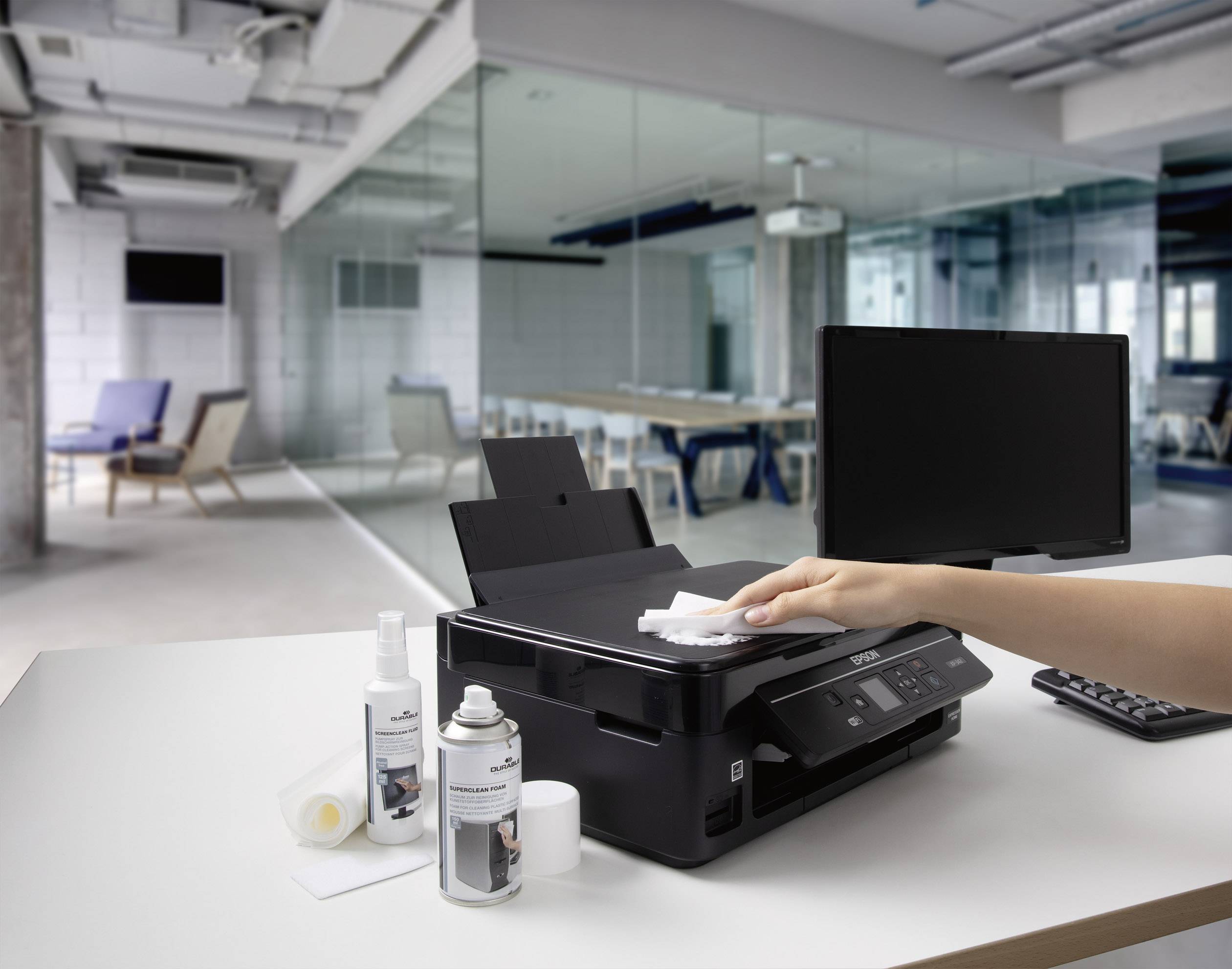 Une personne nettoie une imprimante dans un bureau moderne. À côté de l'imprimante se trouvent des produits de nettoyage, un écran d'ordinateur et un clavier.