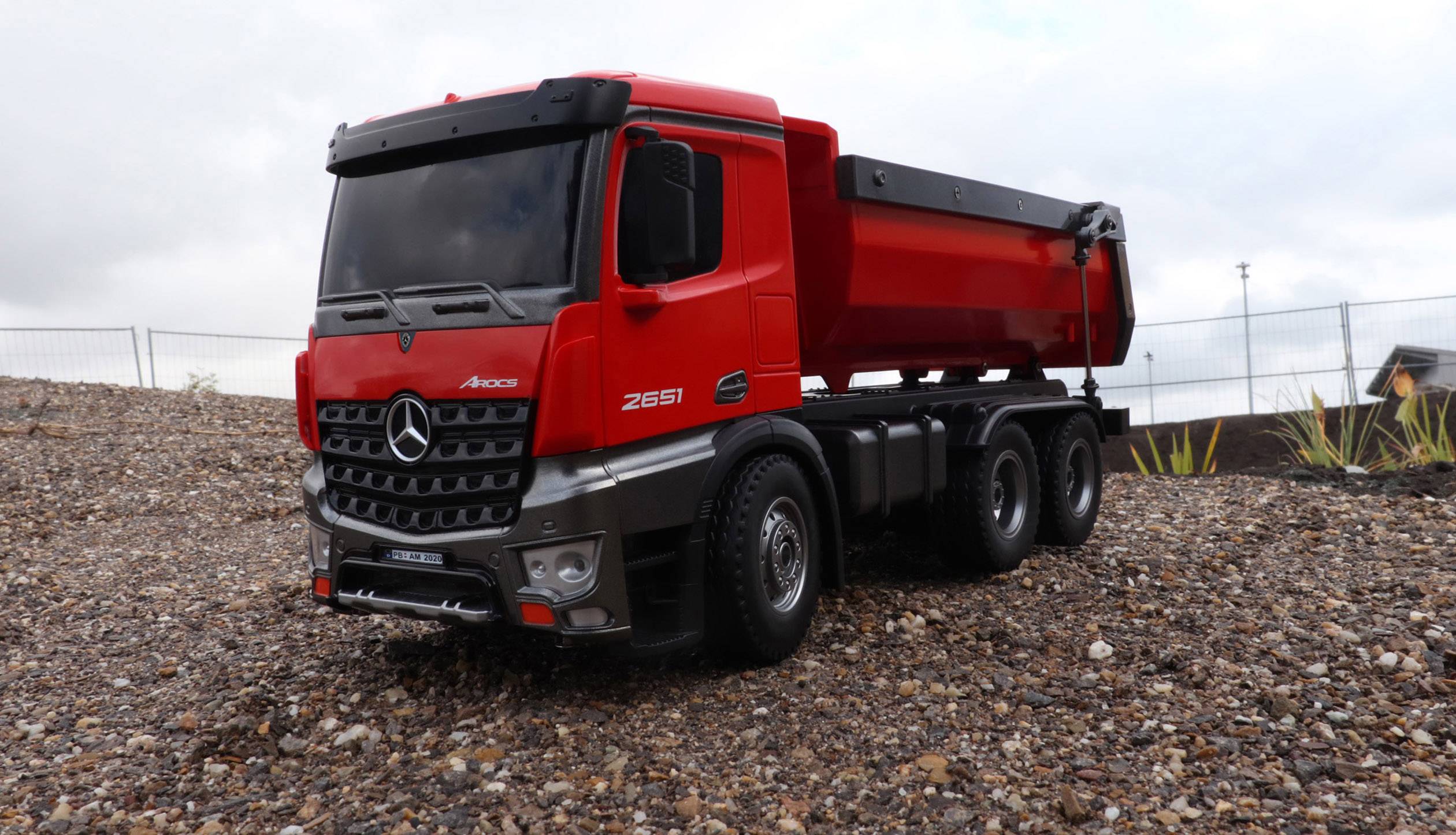 Un camion-model rouge détaillé est stationné sur une surface de gravier. Le ciel est nuageux, et des clôtures sont visibles en arrière-plan.