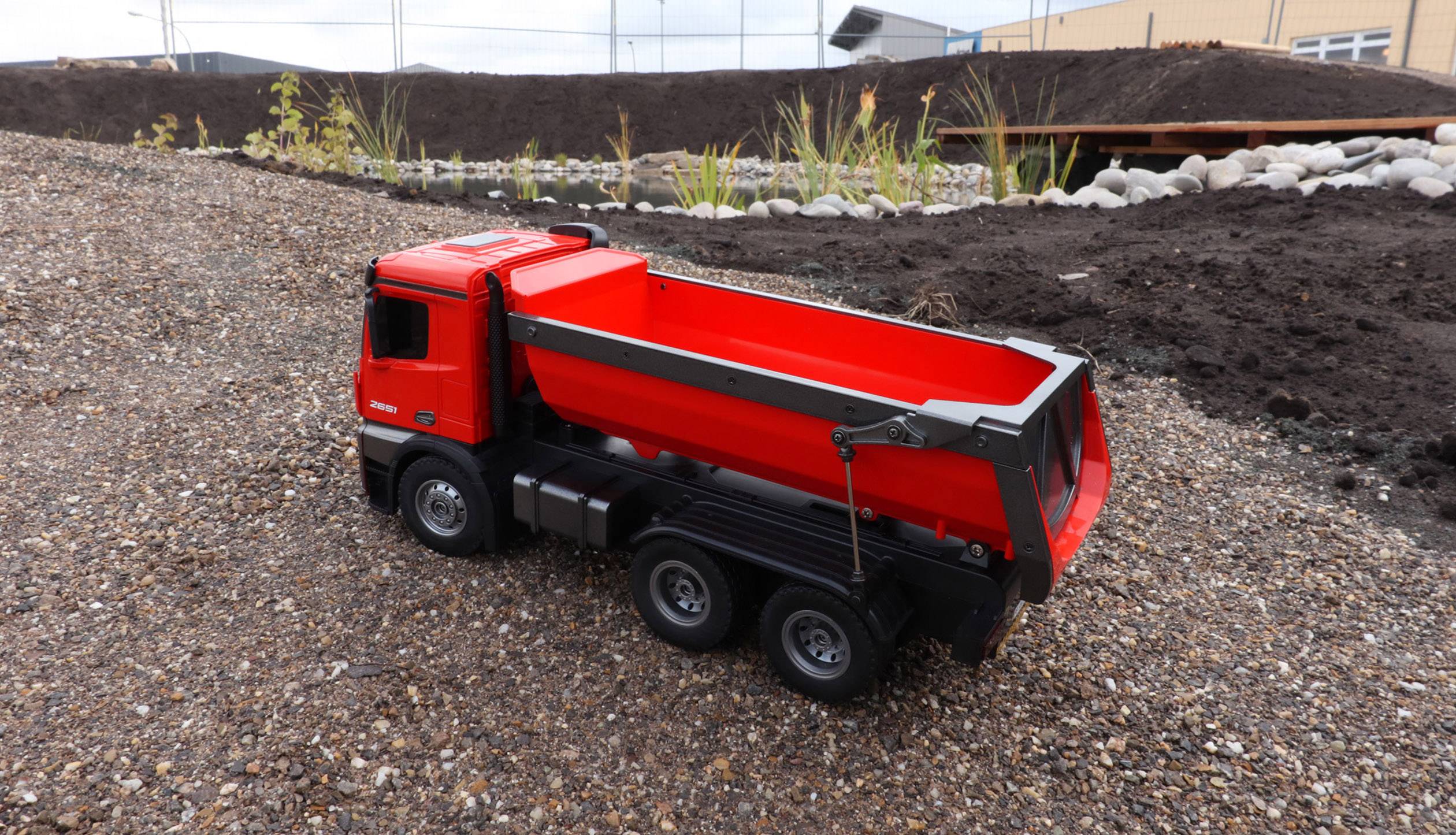 Un camion-jouet rouge est posé sur un sol de gravier devant un talus. Des plantes et un bâtiment sont visibles en arrière-plan.