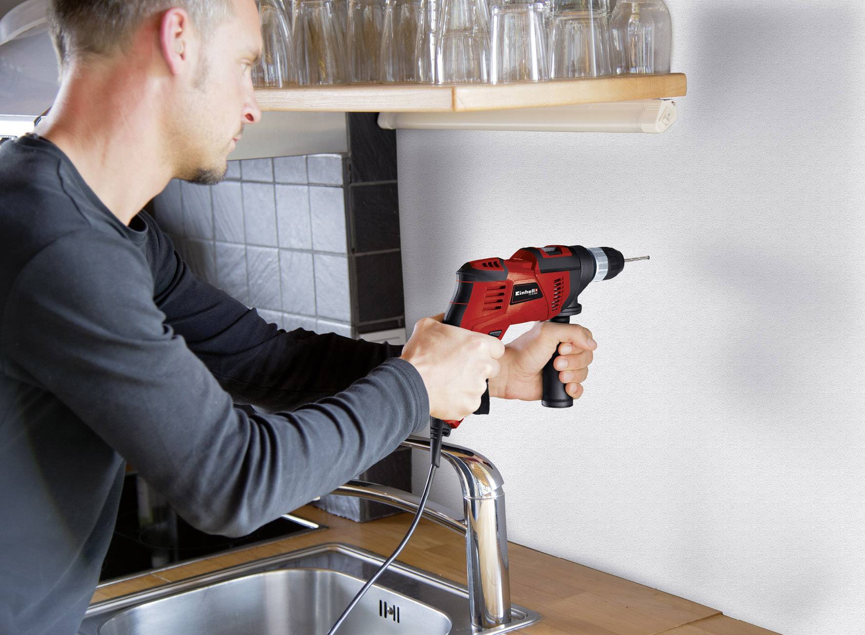 Un homme fore un trou dans le mur au-dessus d'un évier de cuisine à l'aide d'une perceuse rouge.
