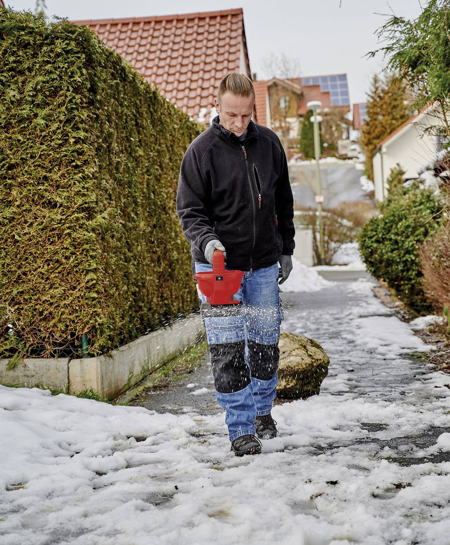 Une personne répand du sel sur un trottoir enneigé devant une maison avec une haie. D'autres maisons sont visibles en arrière-plan.