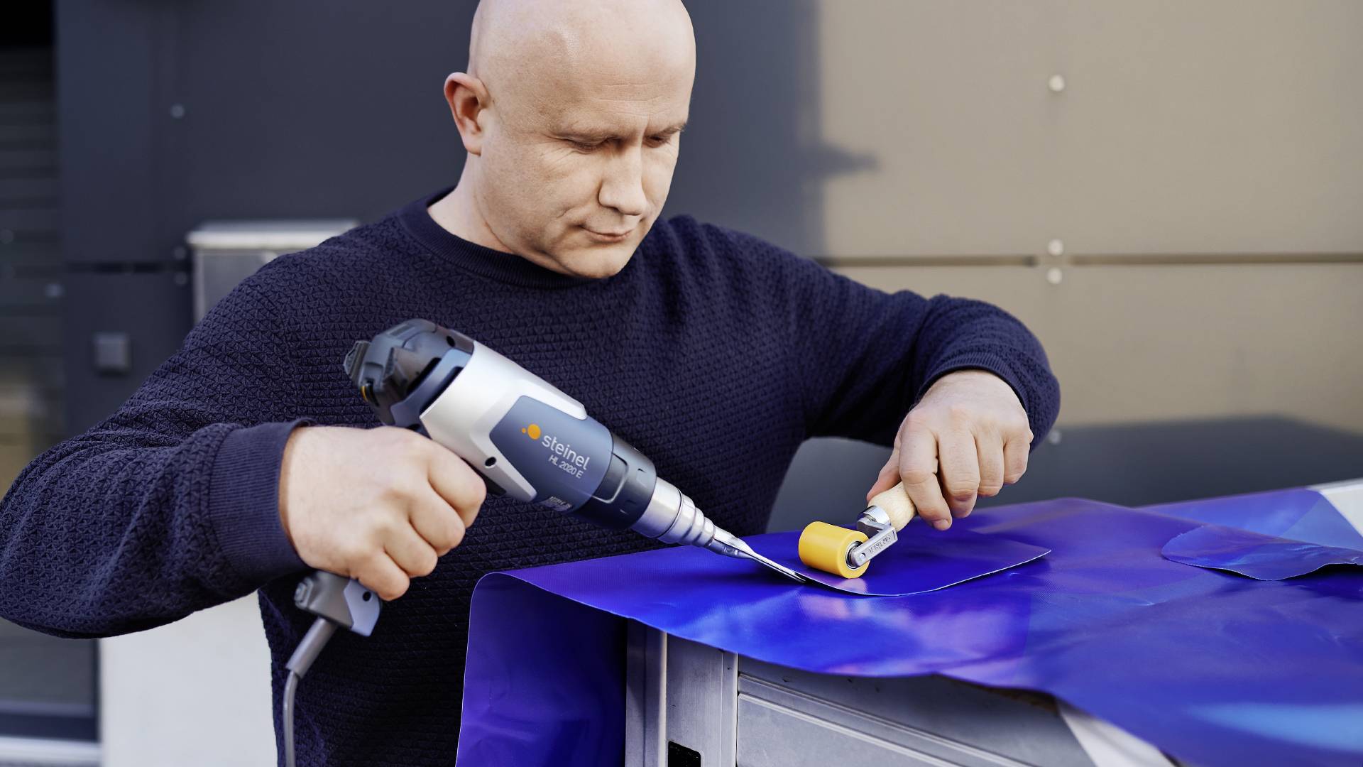 Une personne travaille une feuille bleue à l'aide d'un outil électrique et d'un petit rouleau, probablement dans un contexte artisanal.