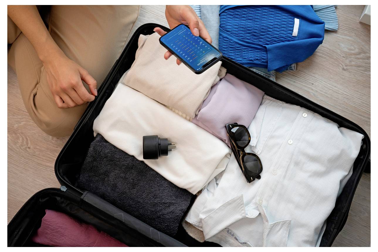 Personne qui range une valise ouverte avec des vêtements pliés, des lunettes de soleil et un téléphone, se préparant pour un voyage.