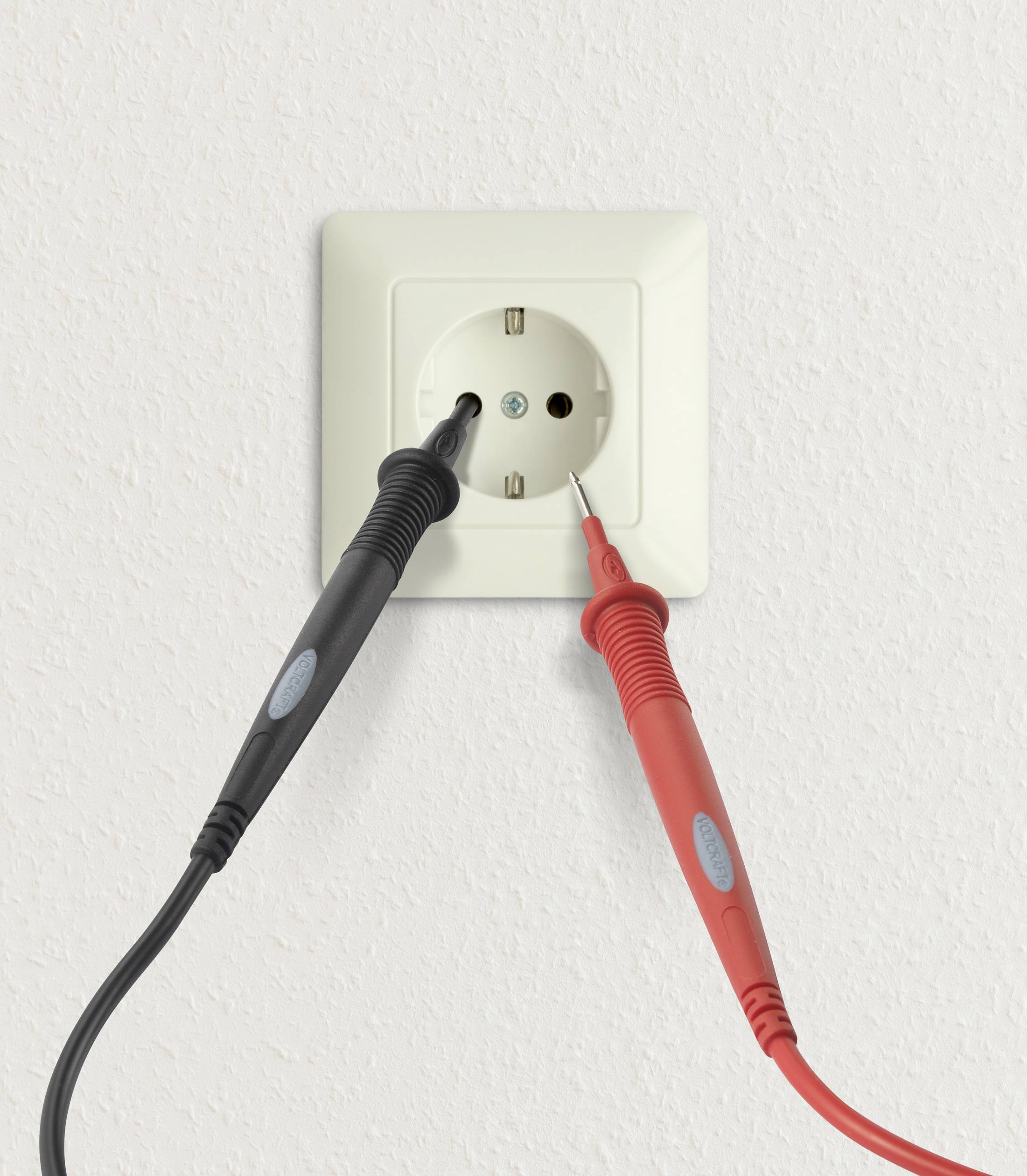 Un ampèremètre avec des câbles rouges et noirs mesure la tension sur une prise électrique blanche dans le mur.