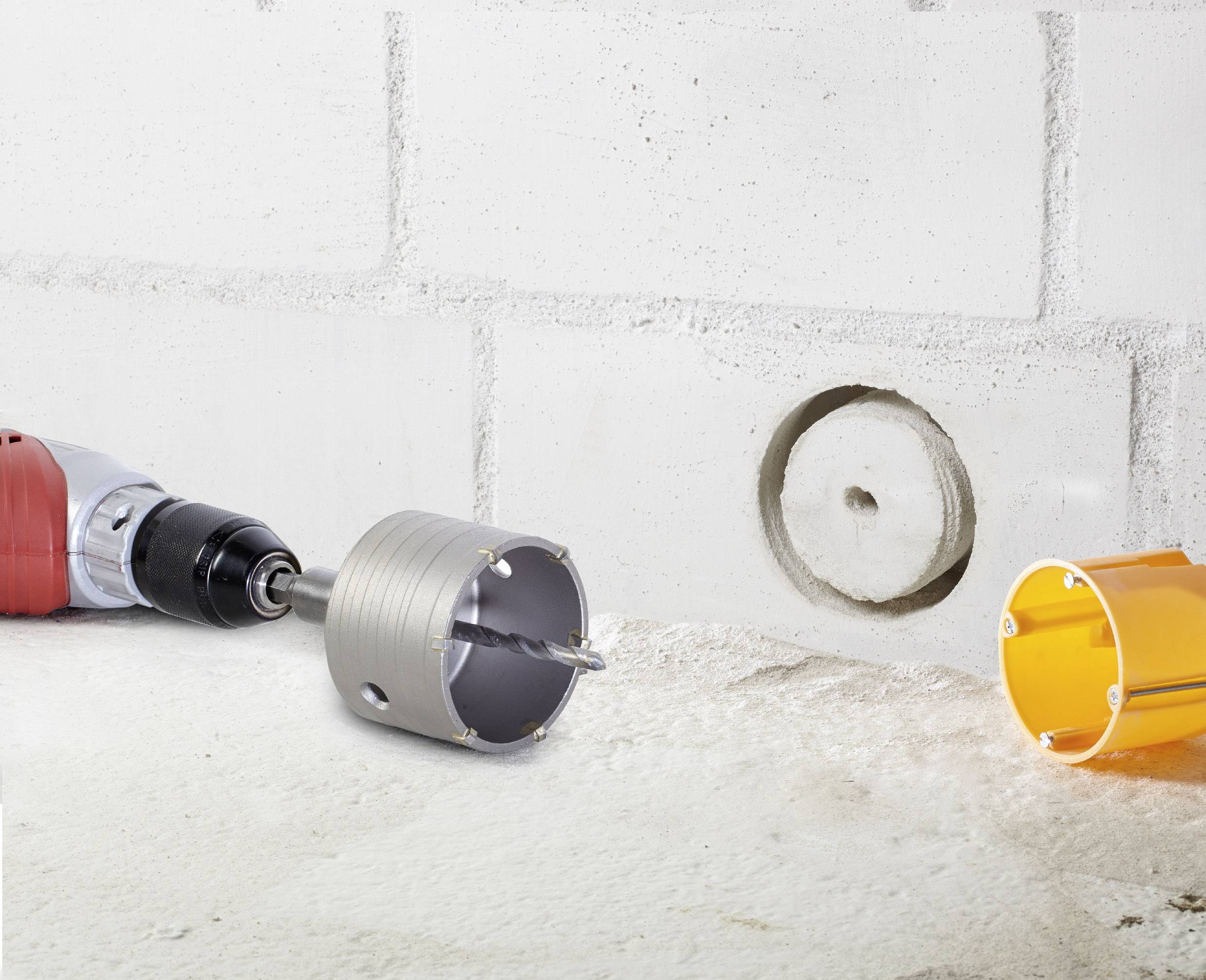 Perceuse avec un accessoire de scie-cloche forant un trou circulaire dans un mur de briques blanches. Un accessoire de scie-cloche orange est posé à côté.