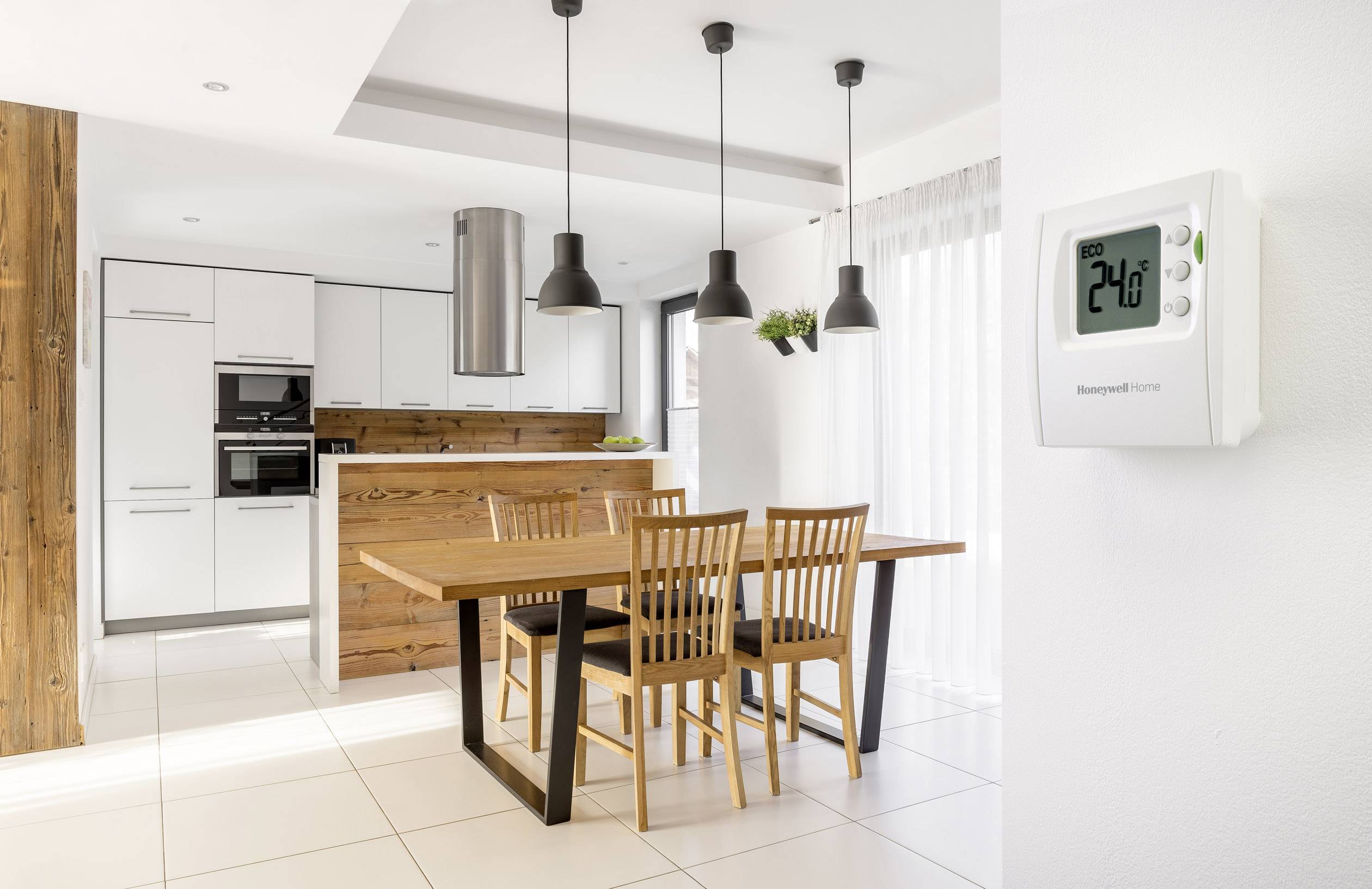Salon lumineux et moderne avec table à manger et chaises, attenant à une cuisine ouverte. Un thermostat est accroché au mur, affichant 24°C.