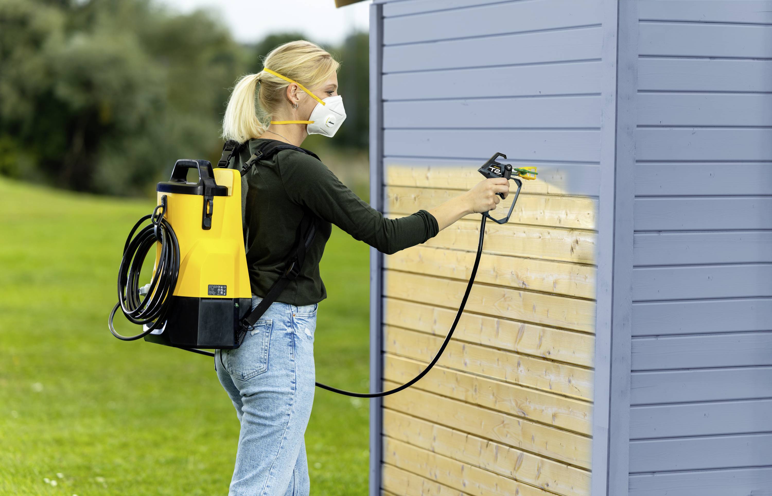 Une personne pulvérise de la peinture sur une surface en bois d'un hangar à l'aide d'un pulvérisateur dorsal jaune. Elle porte un masque de protection respiratoire.