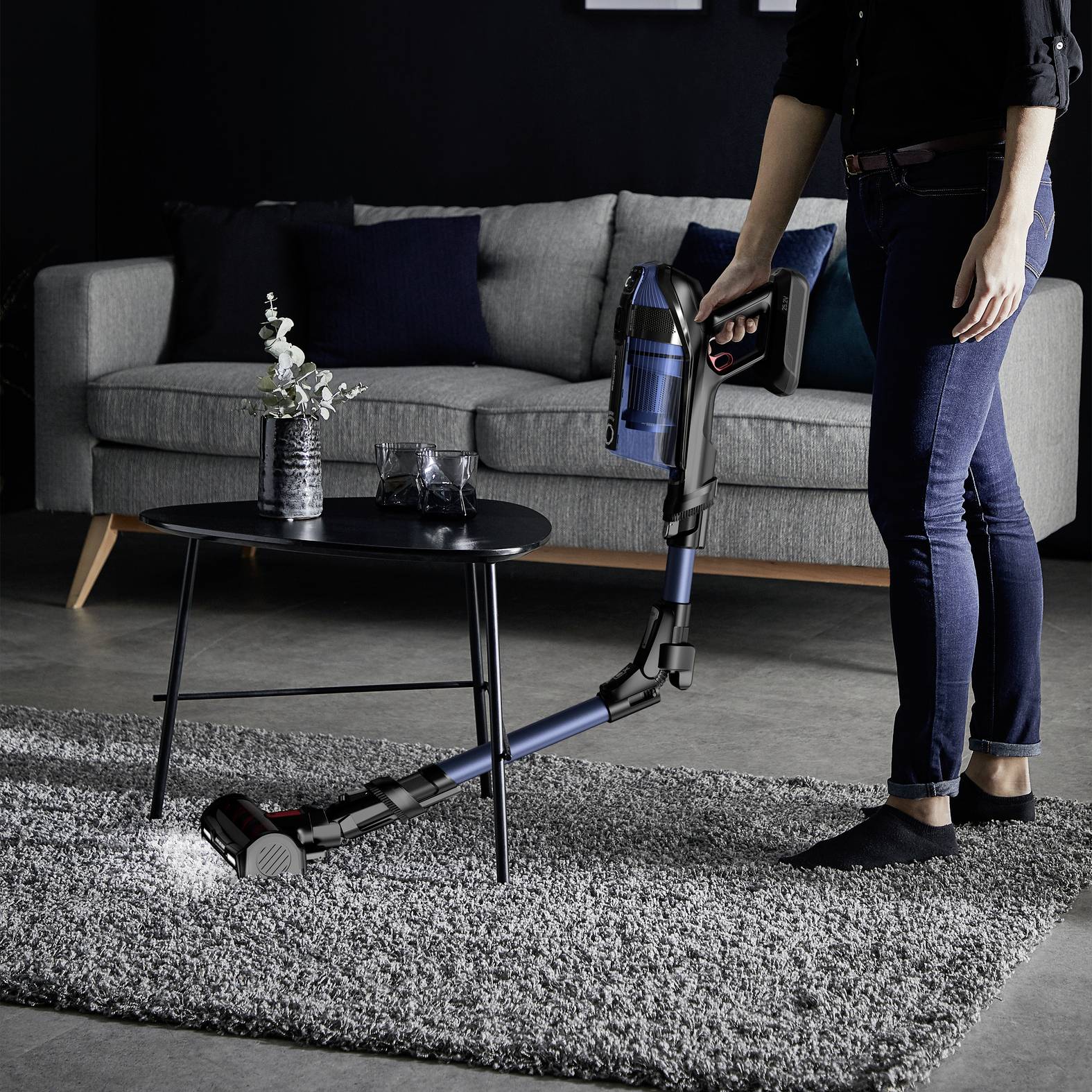 Une personne utilise un aspirateur sans fil sur un tapis dans un salon moderne avec un canapé et une table basse.
