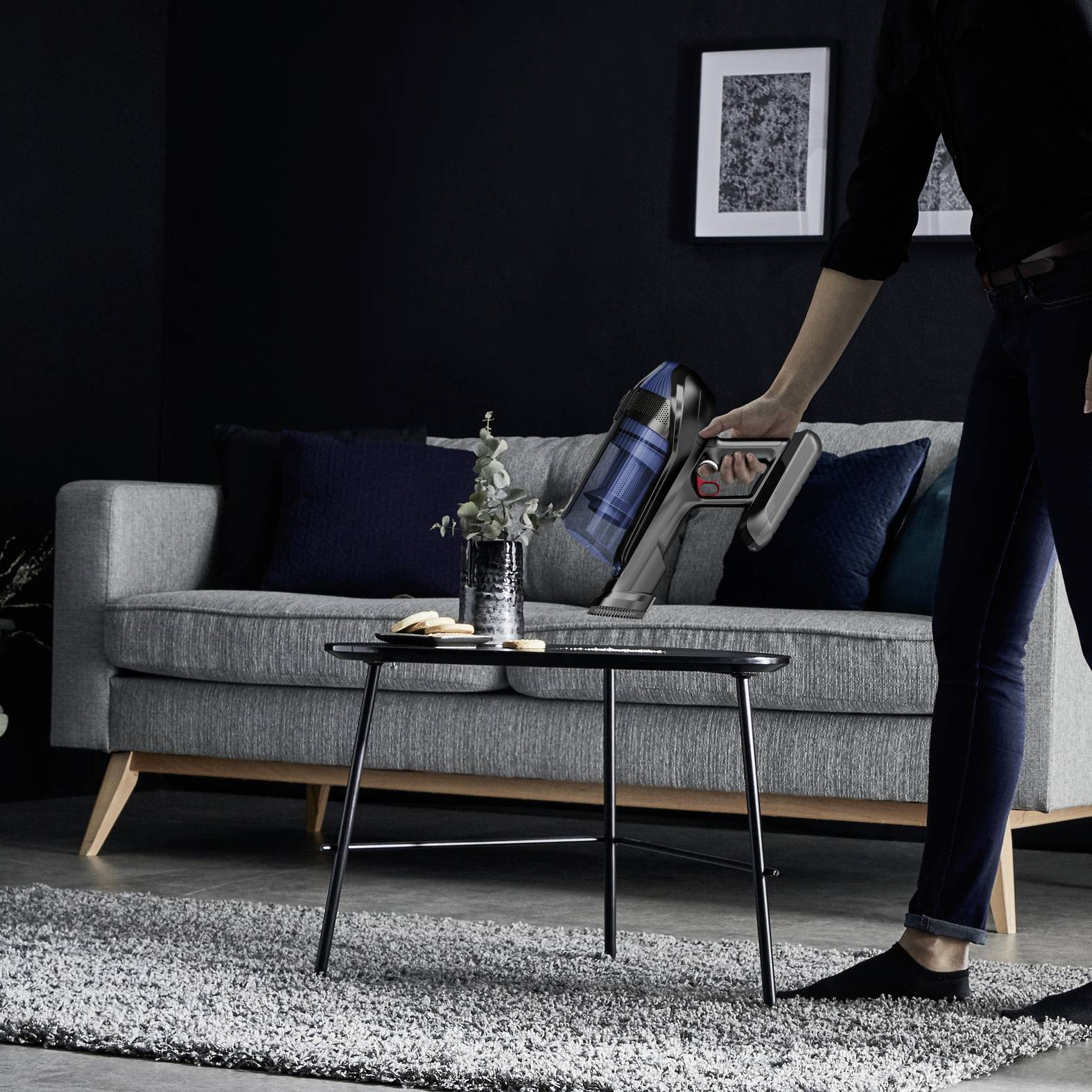 Une personne aspire le tapis dans un salon avec un aspirateur sans fil, tandis qu'un chat est assis sur un canapé gris.