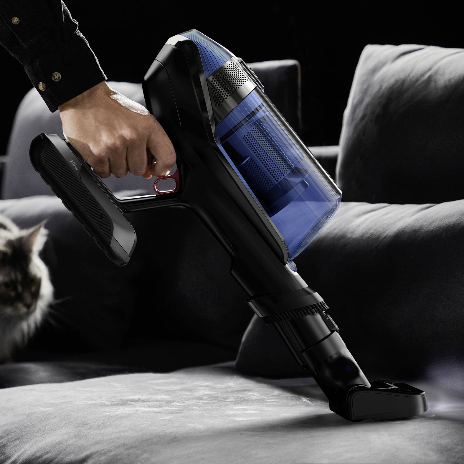 Une personne aspire un canapé gris avec un aspirateur sans fil. Un chat est assis en arrière-plan.