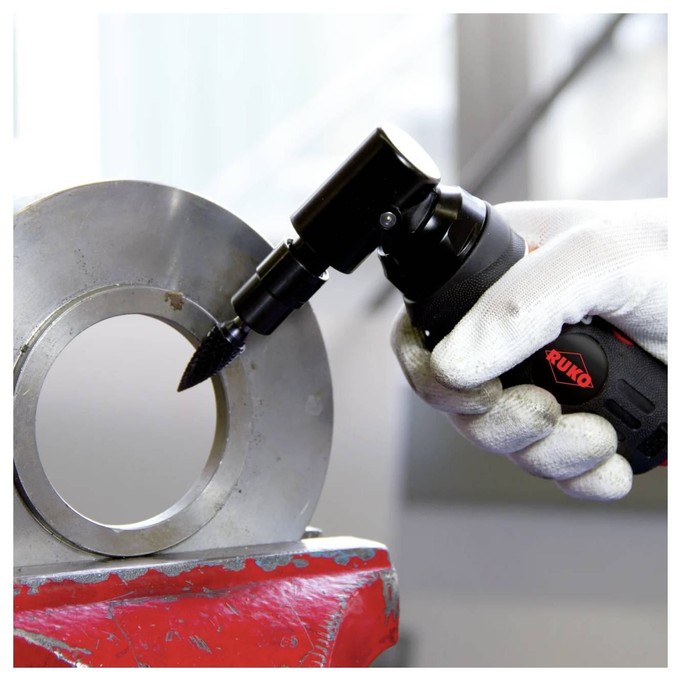 Main gantée utilisant un outil de meulage pneumatique noir sur une grande bague en métal maintenue dans un étau rouge.