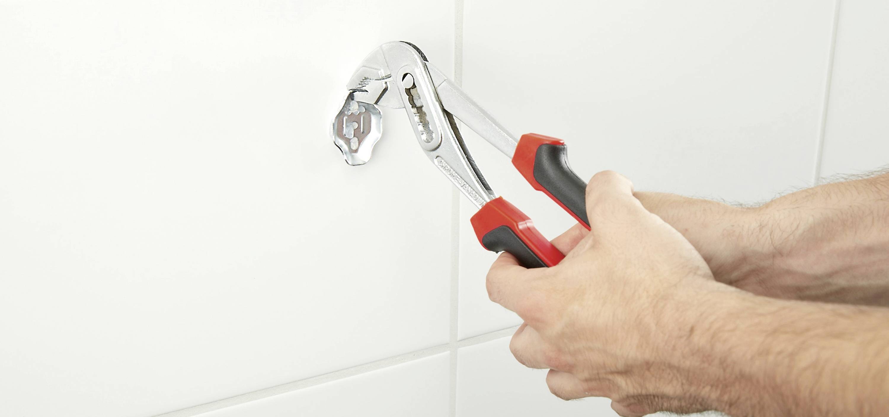 Une personne serre un écrou à molette sur un robinet contre des carreaux blancs, probablement dans une salle de bain ou une cuisine, pour une réparation ou une installation.