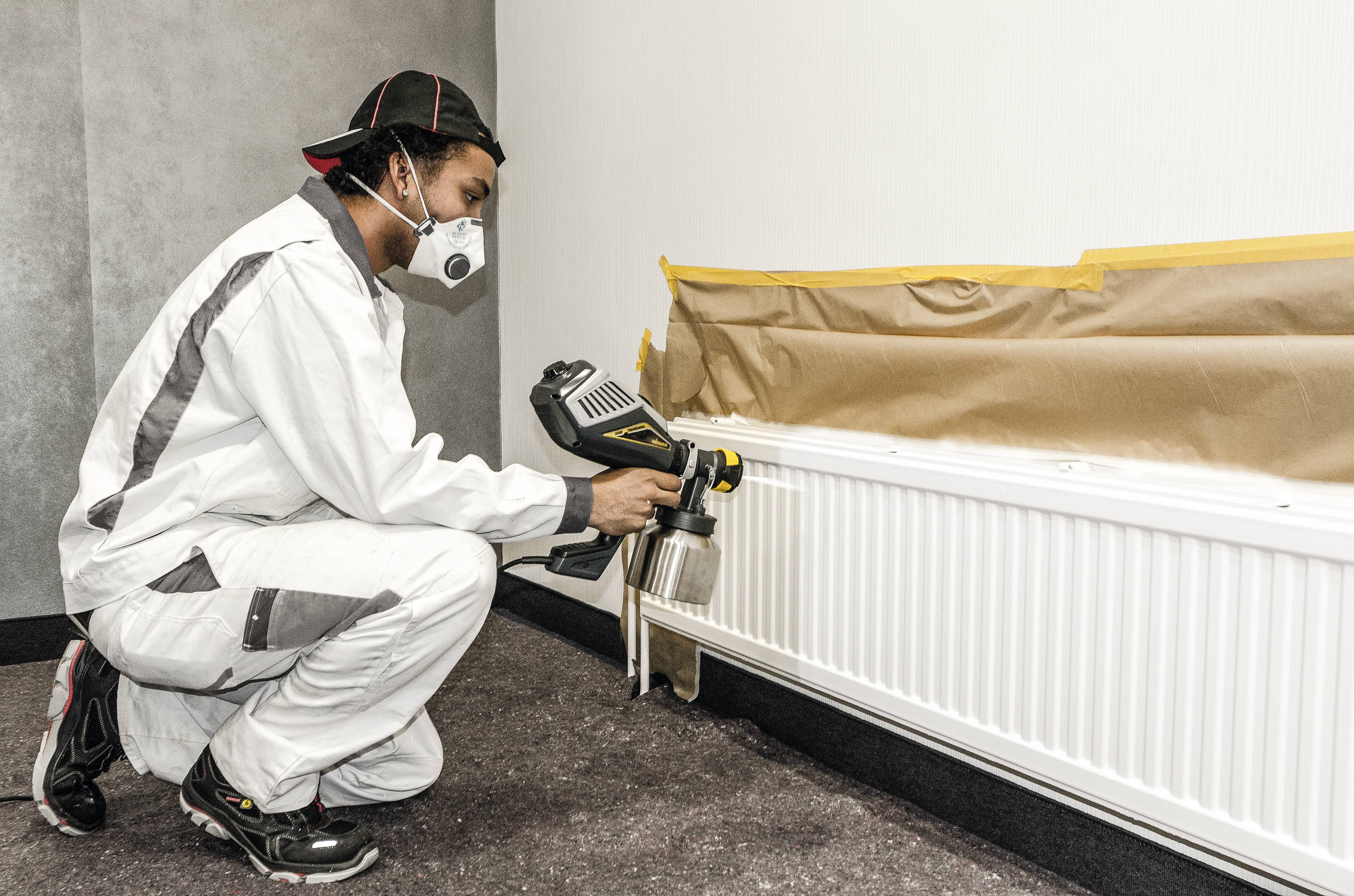 Une personne en tenue de protection pulvérise de la peinture blanche sur un radiateur à l'aide d'un pistolet de peinture. La zone est recouverte de papier.