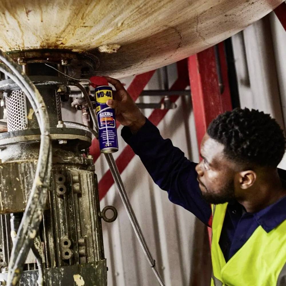 Un ouvrier pulvérise de l'huile sur un grand boulon de machine pour en améliorer le fonctionnement. Il porte un gilet de sécurité.