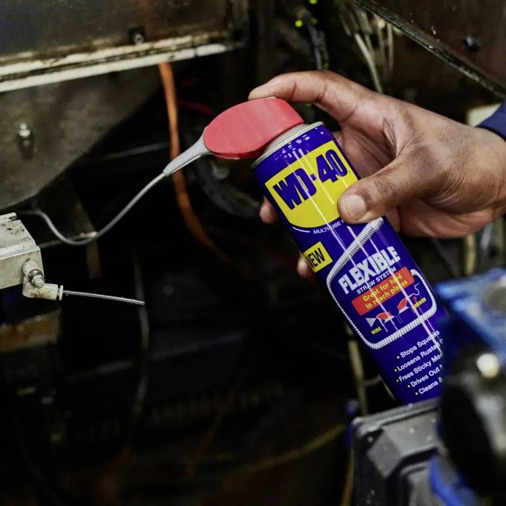 Une main tient un aérosol WD-40 FLEXIBLE avec un tube de pulvérisation flexible. La personne est en train d'appliquer le produit sur un composant de machine.