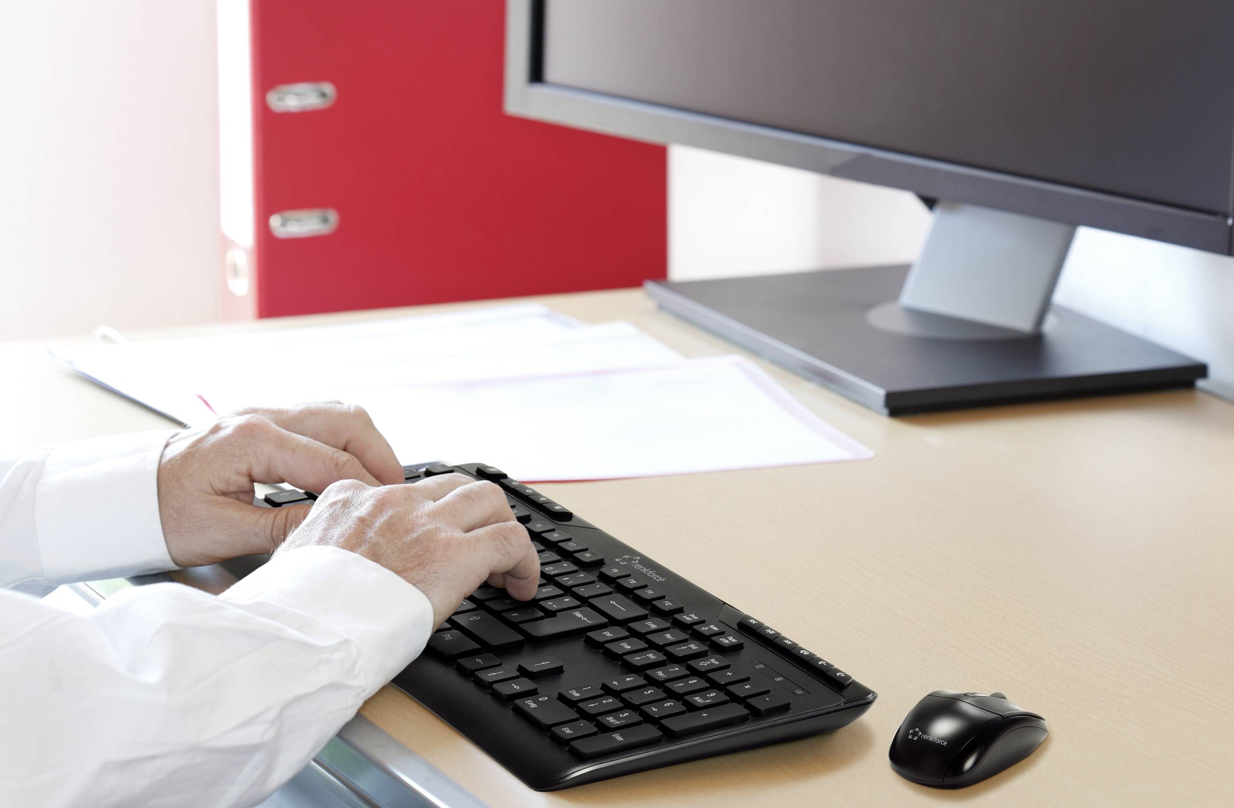 Une personne tape sur un clavier d'ordinateur devant un écran, avec un dossier rouge en arrière-plan sur un bureau.