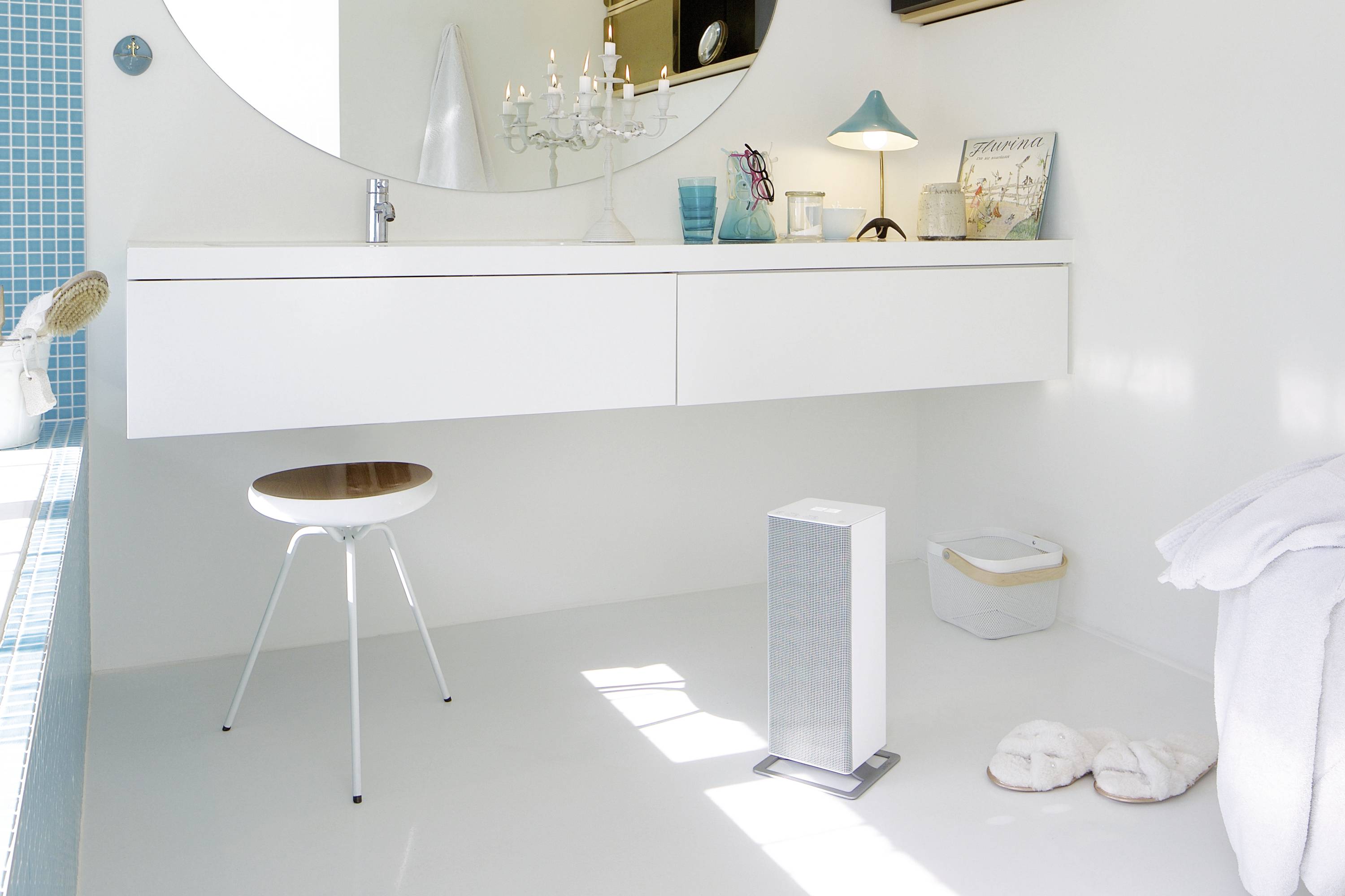 Salle de bain moderne et blanche avec un miroir rond, un lavabo suspendu et un tabouret. Une lampe et des livres sont posés sur le plan de toilette.