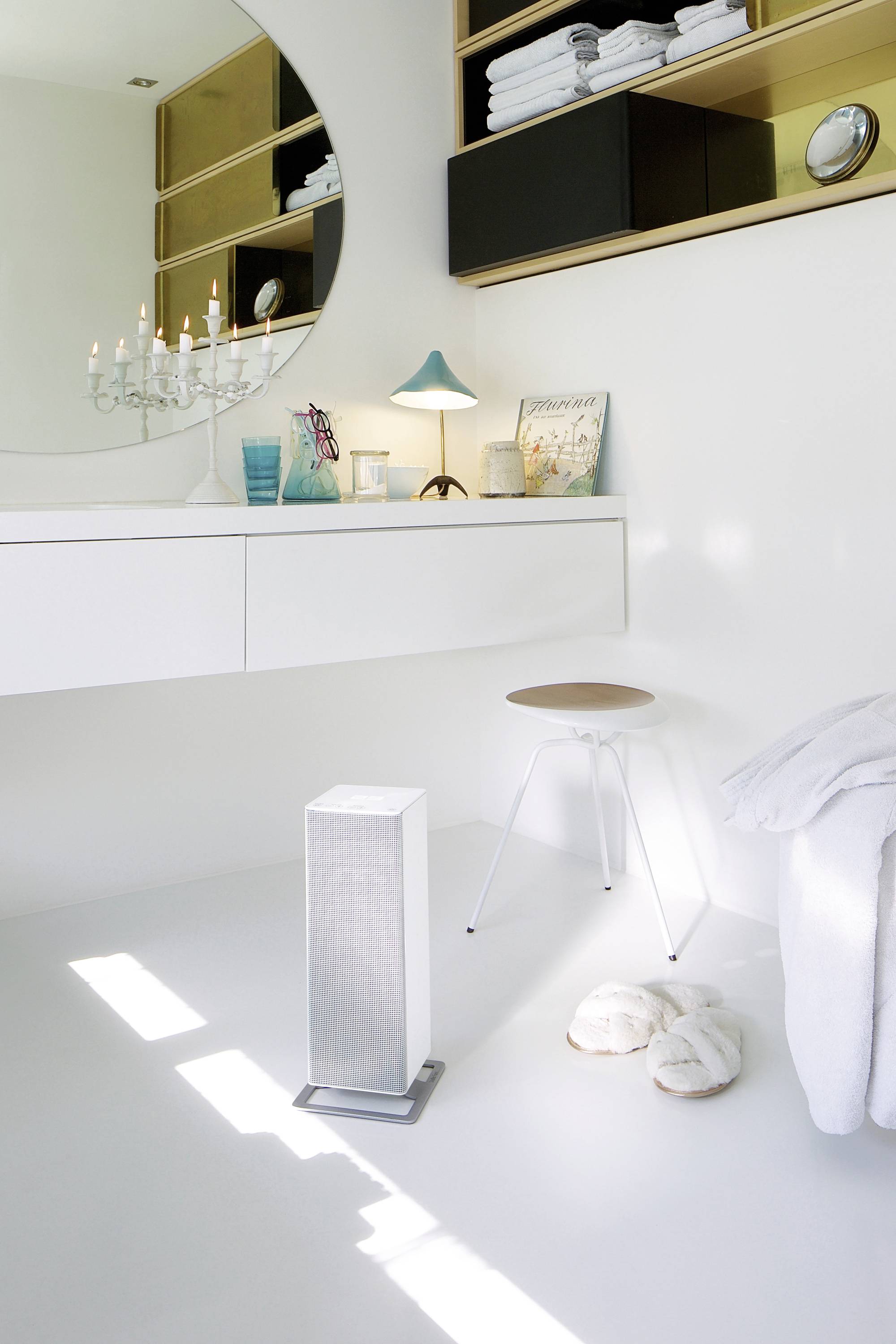 Salle de bain lumineuse avec lavabo blanc, miroir rond, un tabouret et des objets de décoration tels qu'un bougeoir et un abat-jour.
