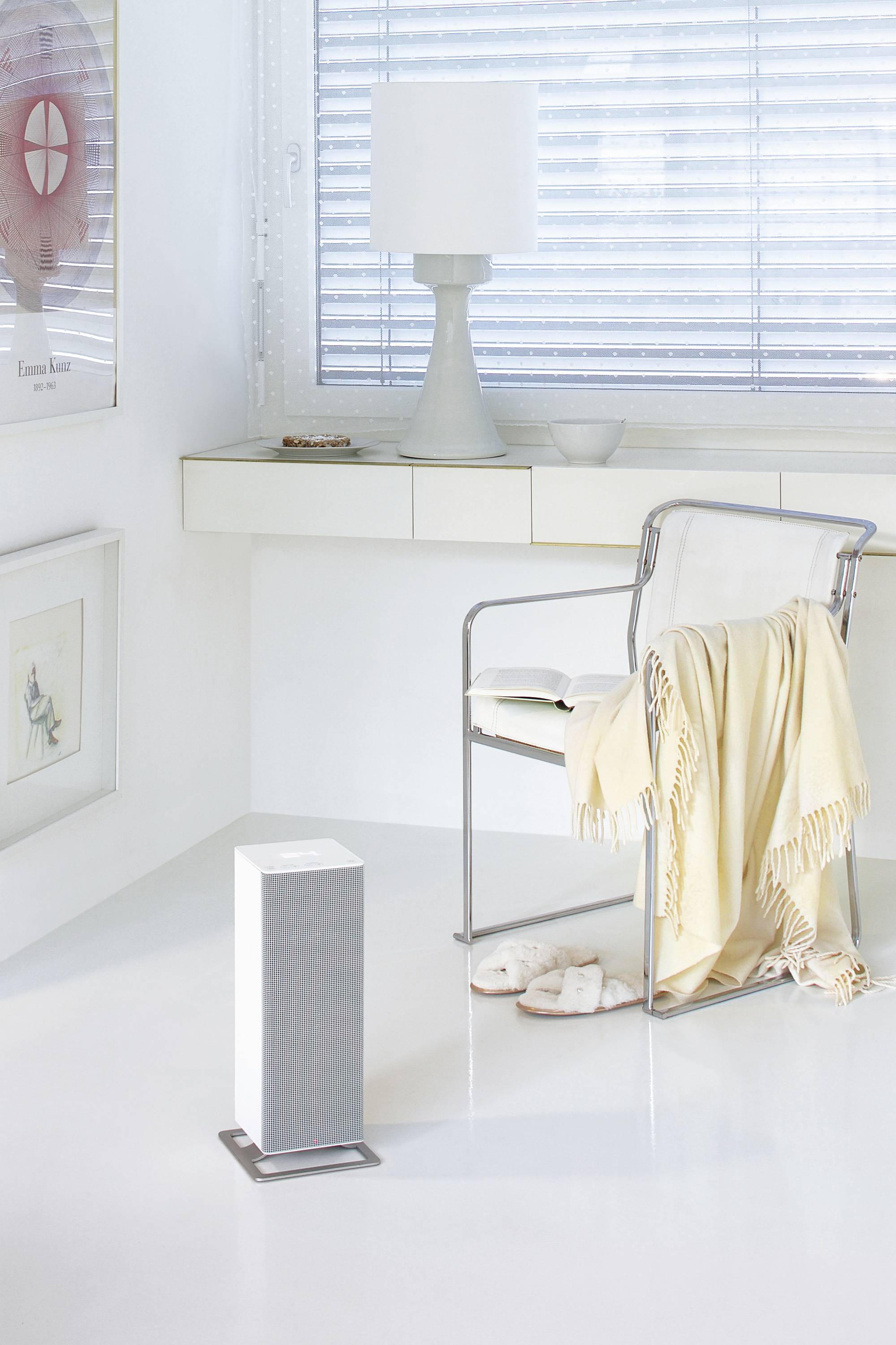 Un espace moderne et lumineux avec une chaise élégante, une lampe et un purificateur d'air à côté d'une fenêtre équipée de stores et décorée d'œuvres d'art.