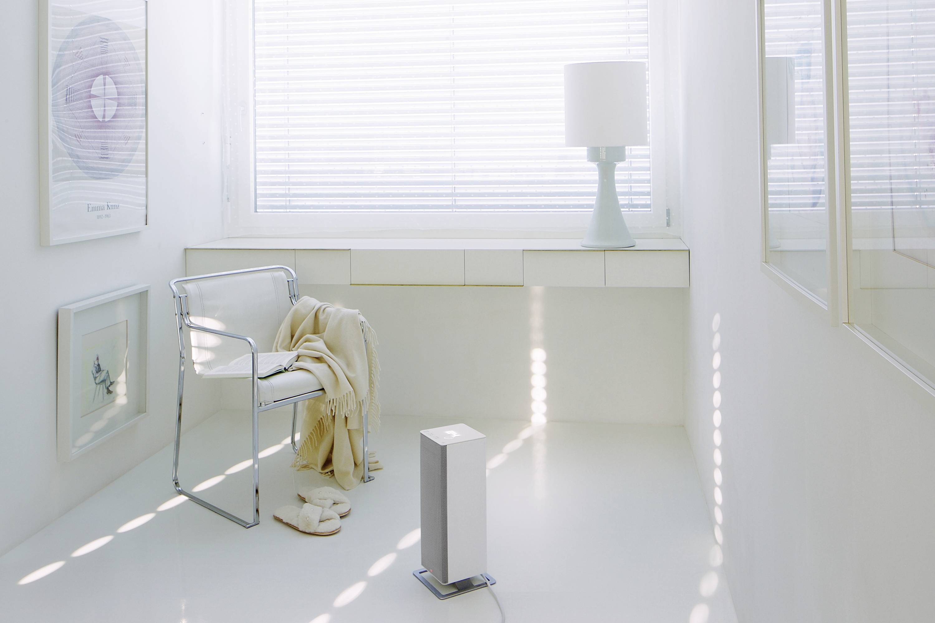 Une pièce lumineuse avec une chaise sur laquelle est posée une couverture, et un petit purificateur d'air debout. À l'arrière-plan, une fenêtre avec des stores.
