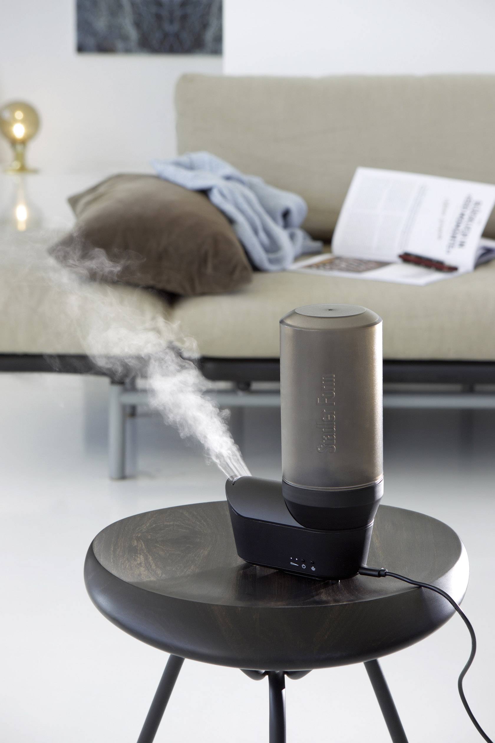 Un humidificateur émet de la vapeur sur une table, avec un canapé et une couverture en arrière-plan, un livre ouvert posé dessus.