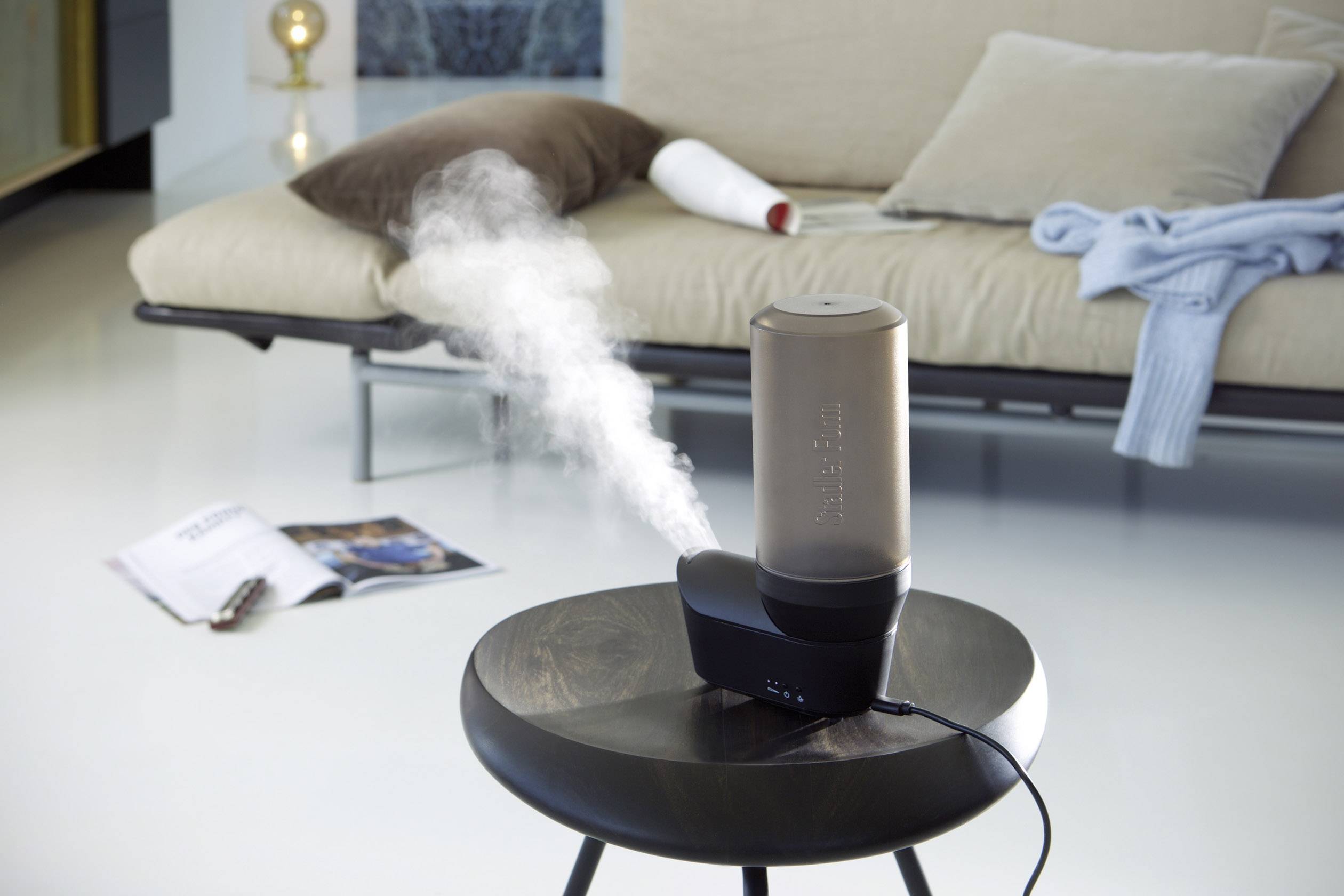 Un humidificateur est posé sur une table et produit de la vapeur. En arrière-plan, on aperçoit un canapé, un coussin et un magazine ouvert.