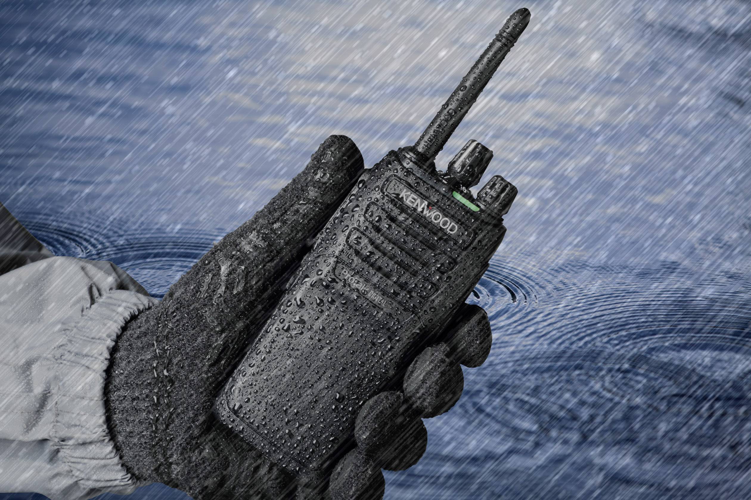 Un talkie-walkie recouvert d'eau est tenu par une main gantée sous la pluie, illustrant sa robustesse et son étanchéité.