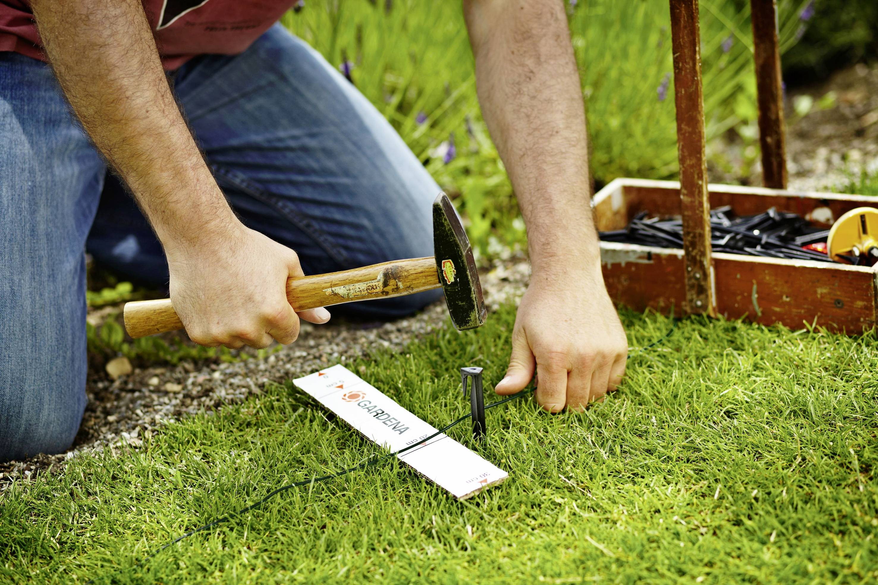 Une personne s'agenouille dans l'herbe et enfonce un clou dans le sol à l'aide d'un marteau. Une boîte à outils est visible en arrière-plan.