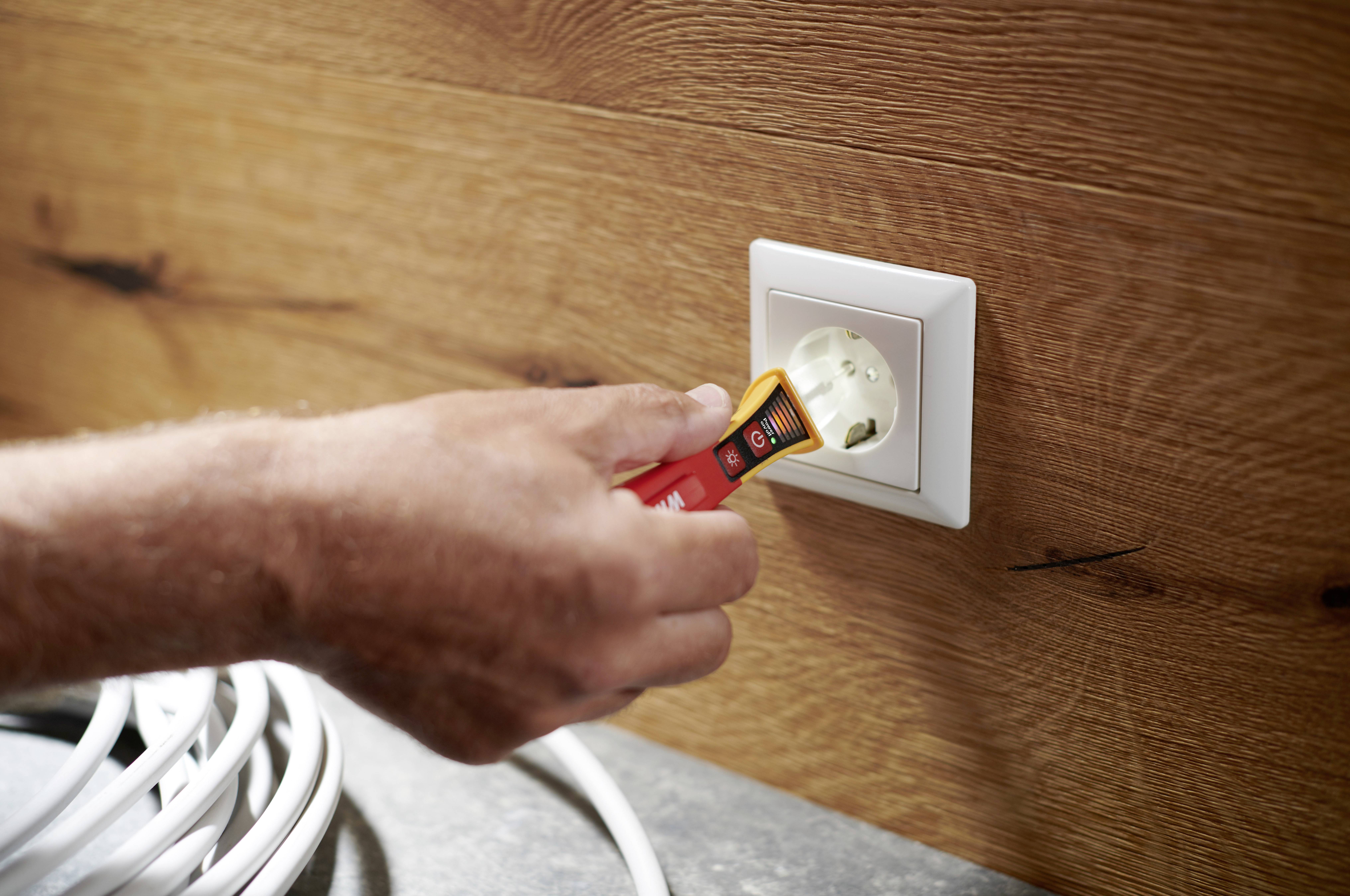 Une main tient un testeur de tension et vérifie une prise électrique blanche sur un lambris en bois. Un câble blanc est posé à côté.