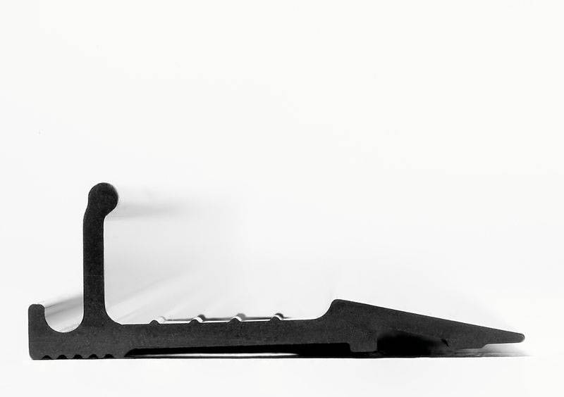 Pièce métallique noire, vue de côté, montrant une structure rectangulaire avec une base dentelée et une extrémité inclinée sur un fond blanc.