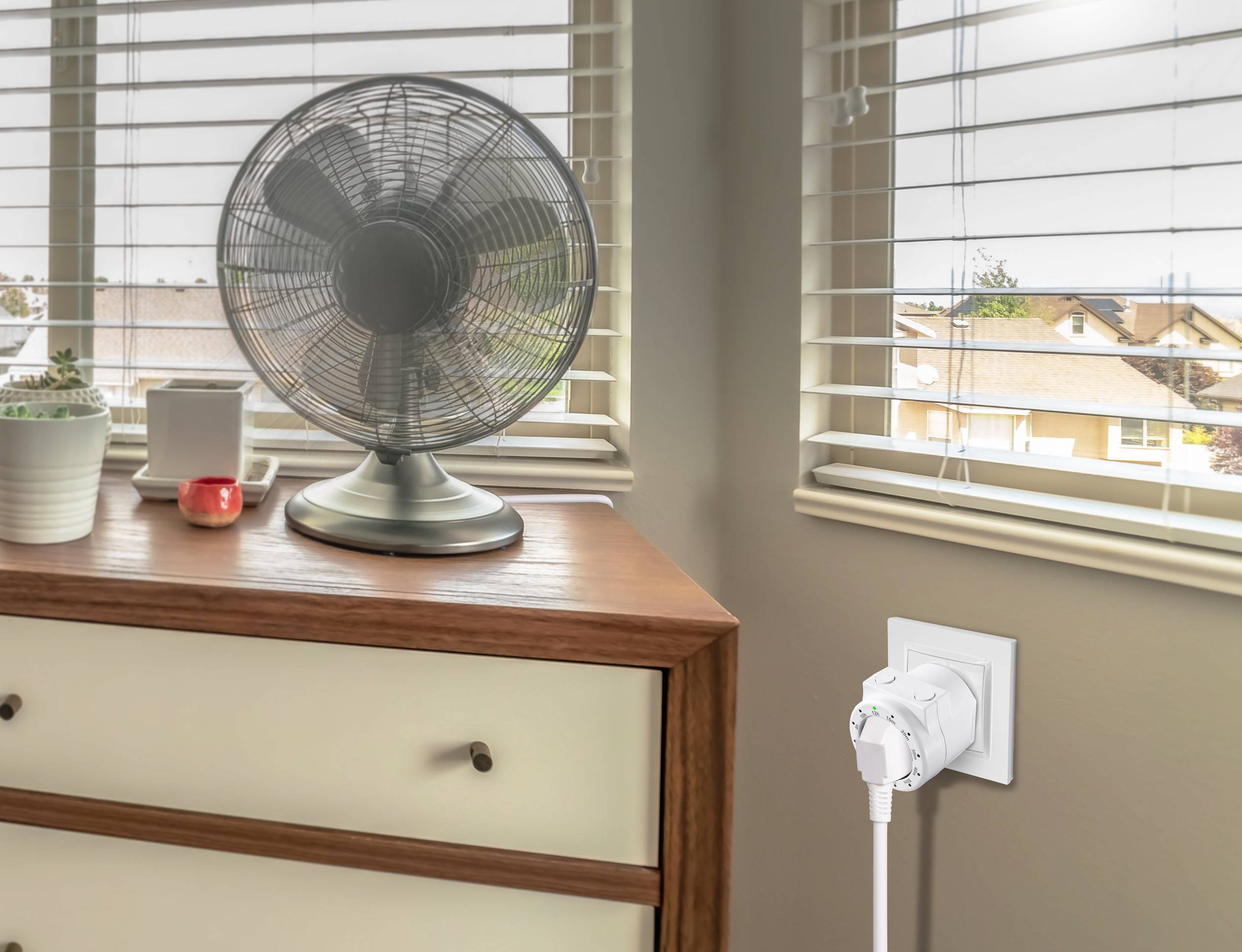 Un ventilateur est posé sur une commode dans une pièce lumineuse. Un appareil domotique est branché à la prise murale.