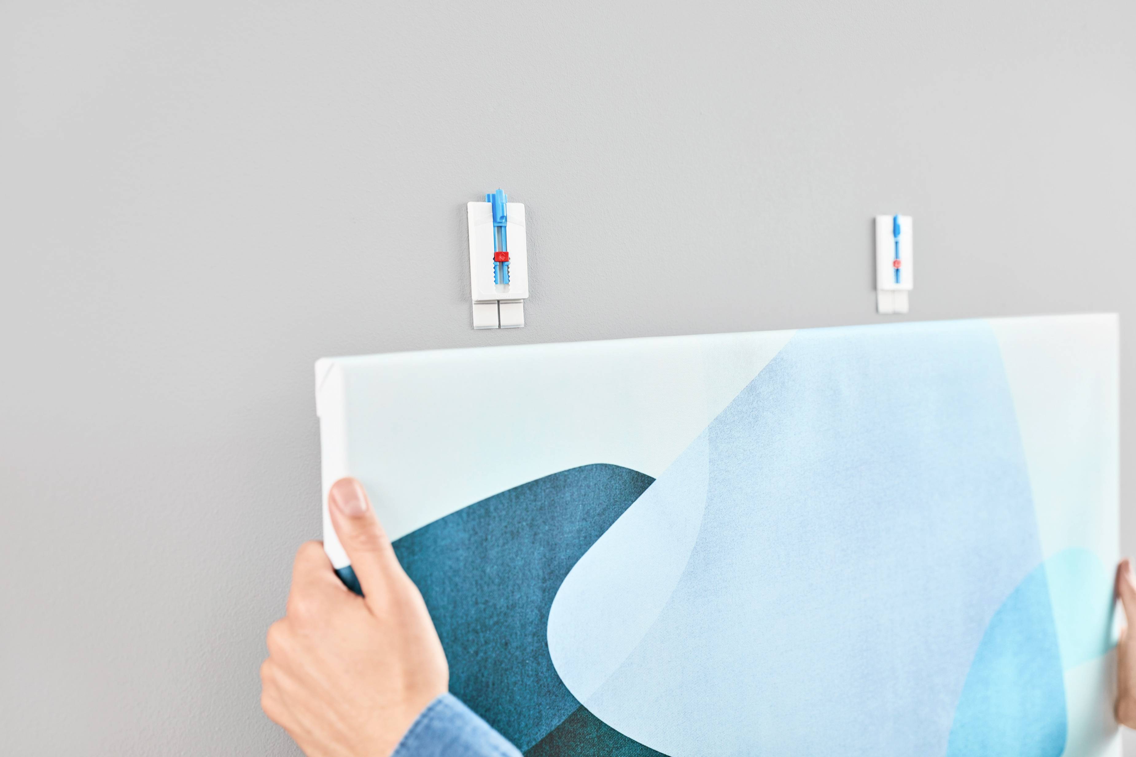 Deux mains accrochent une toile aux formes géométriques sur un mur gris, fixée avec des crochets bleus.