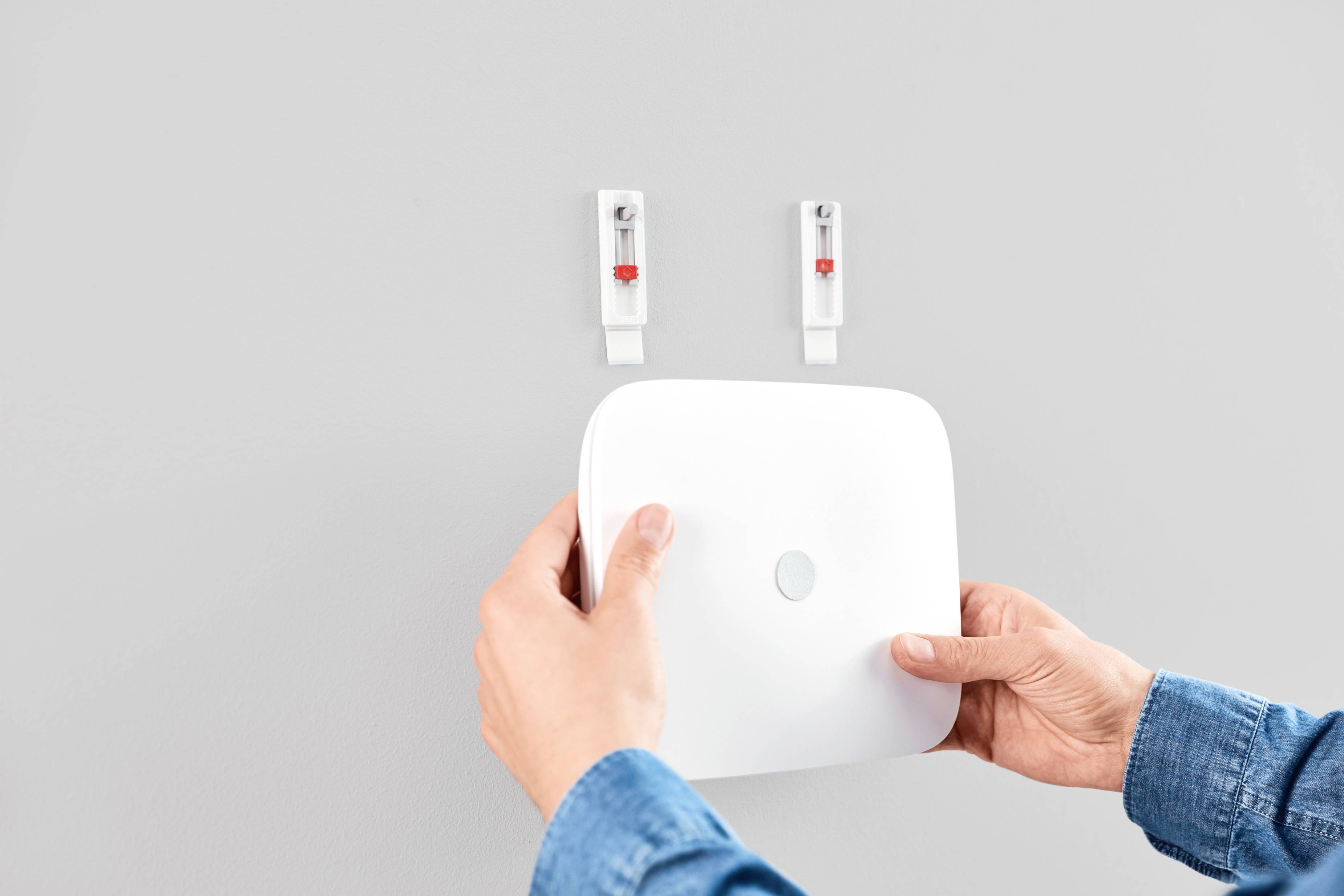 Une personne installe un dispositif carré blanc sur un mur gris. Des mains tiennent l'appareil, qui est positionné entre deux supports muraux.