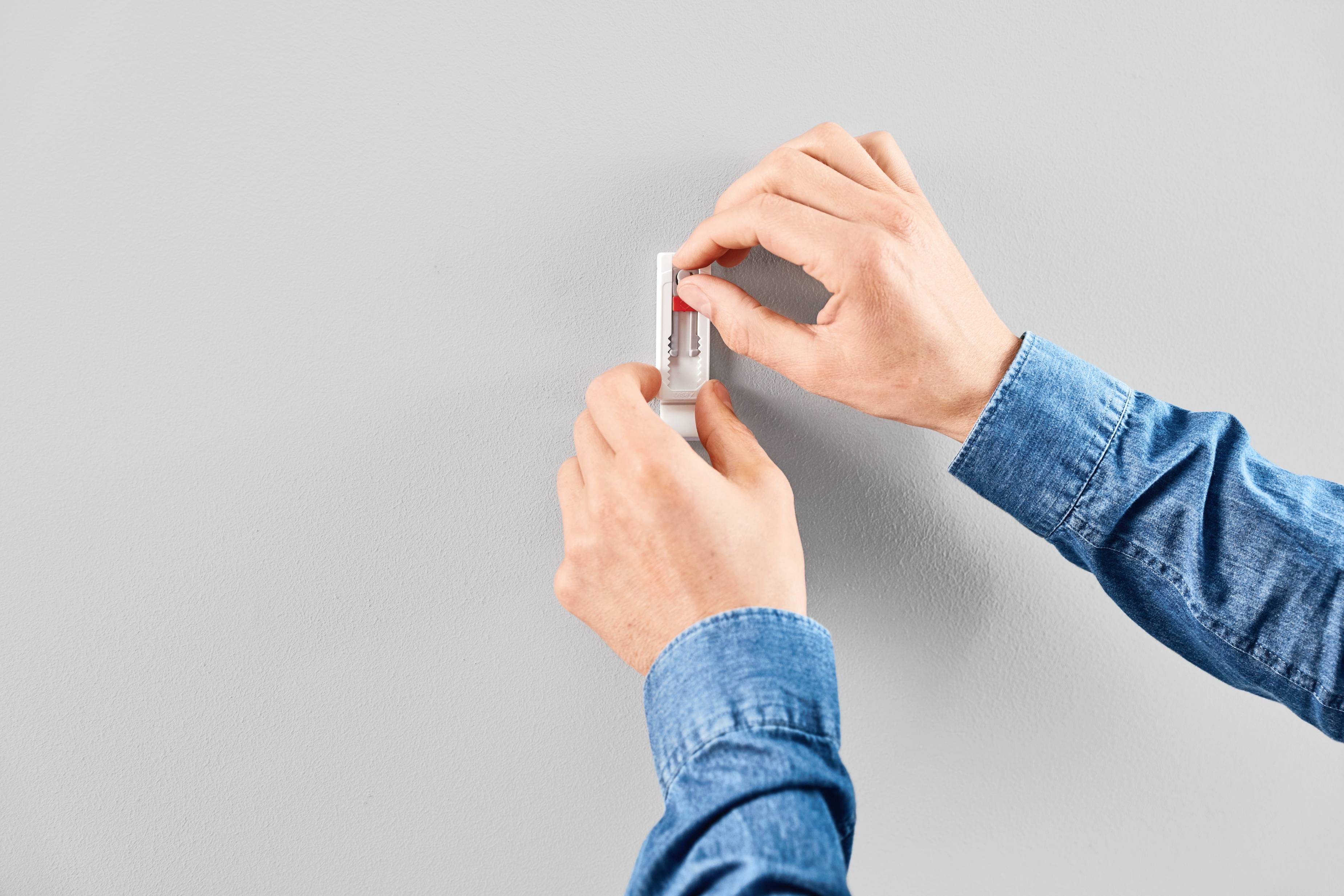 Une personne fixe un petit objet rectangulaire avec un dos adhésif sur un mur gris.