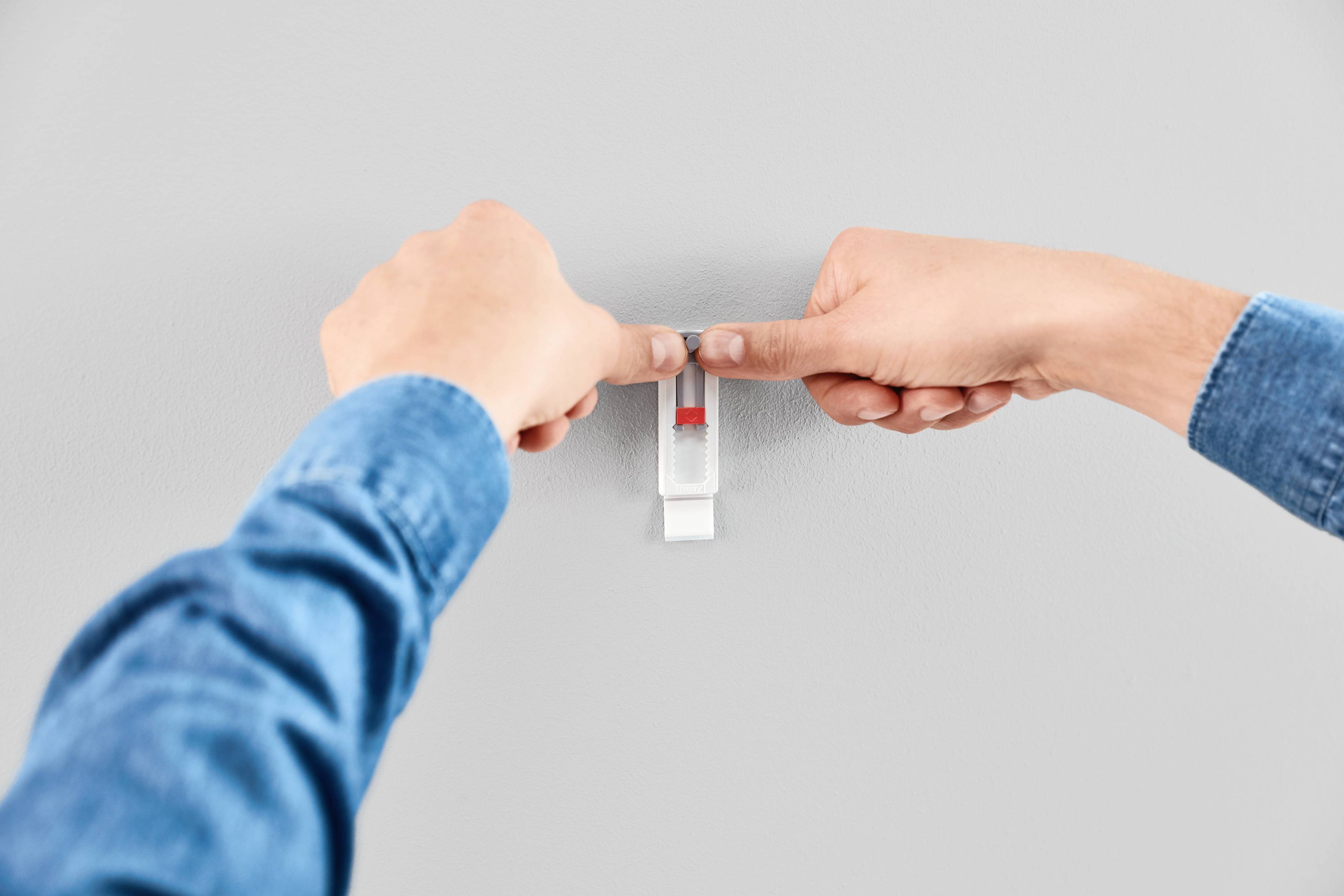 Des mains gantées de bleu accrochent un petit crochet blanc avec un marquage rouge sur un mur lisse et gris.