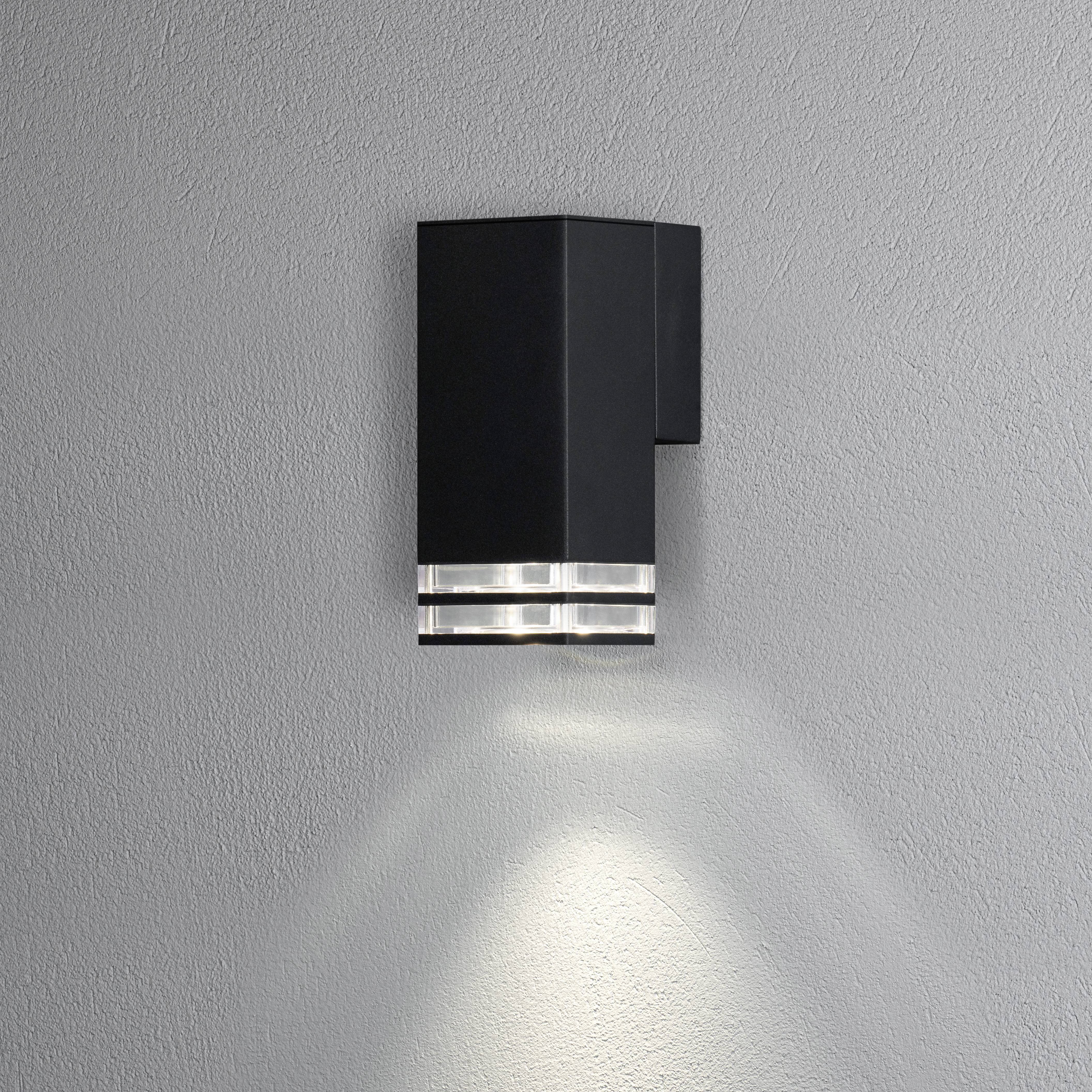 Un luminaire mural moderne en noir diffuse une lumière dirigée vers le bas le long d'un mur gris.