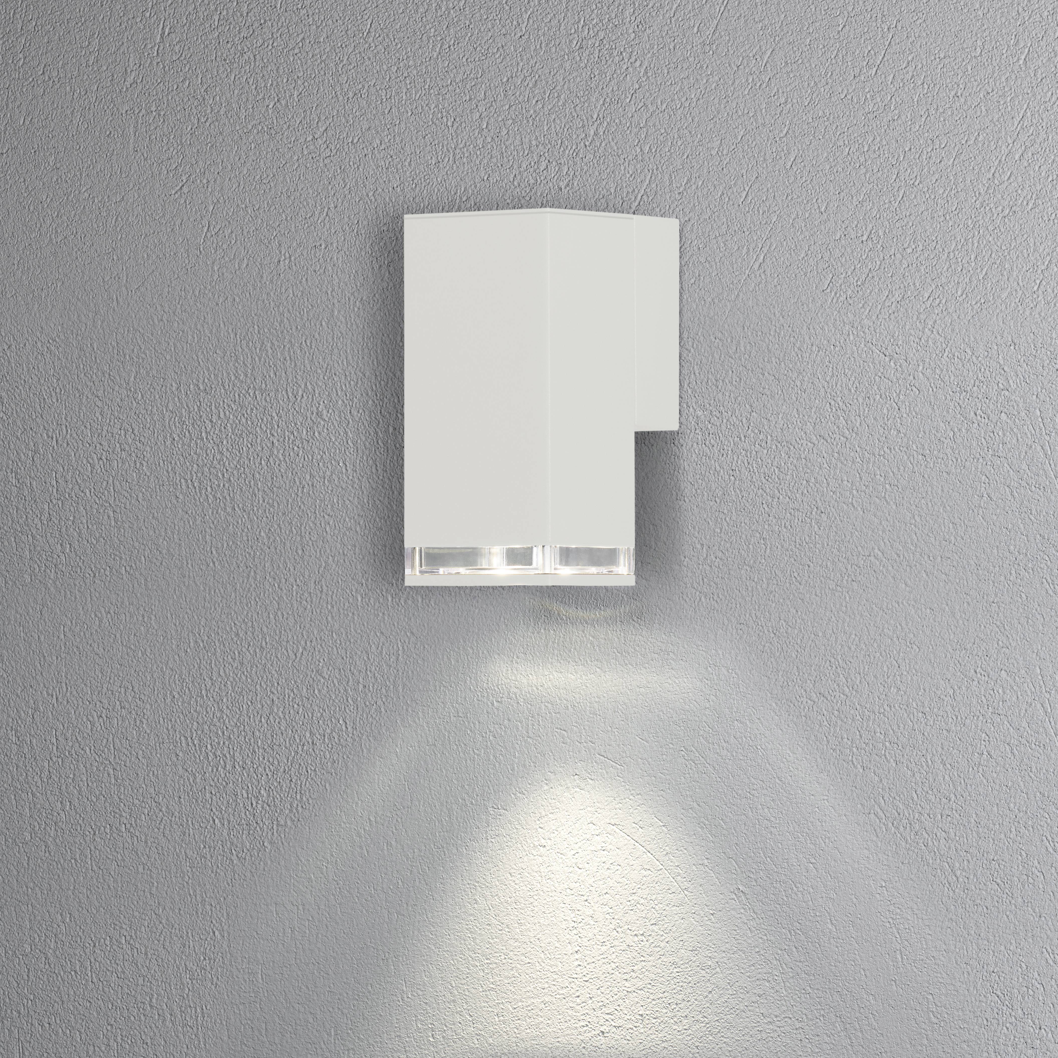 Un applique murale moderne, blanche, au design minimaliste, projetant une lumière douce vers le bas sur un mur gris.