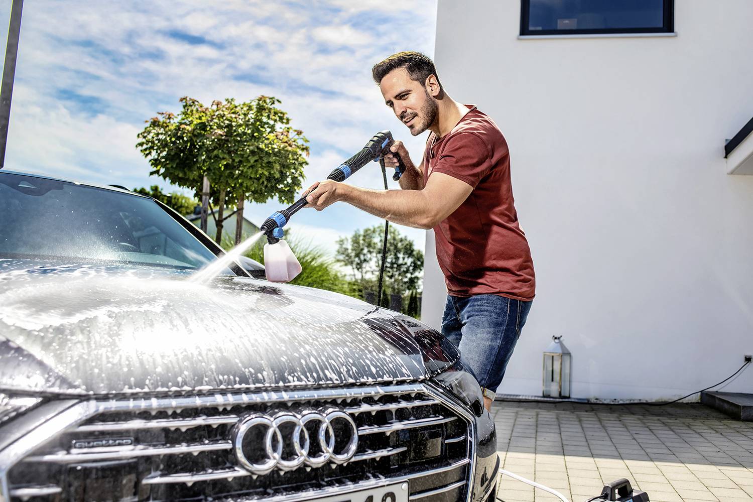 Un homme lave une voiture avec un nettoyeur haute pression sur une allée. Un pavillon moderne est visible en arrière-plan.