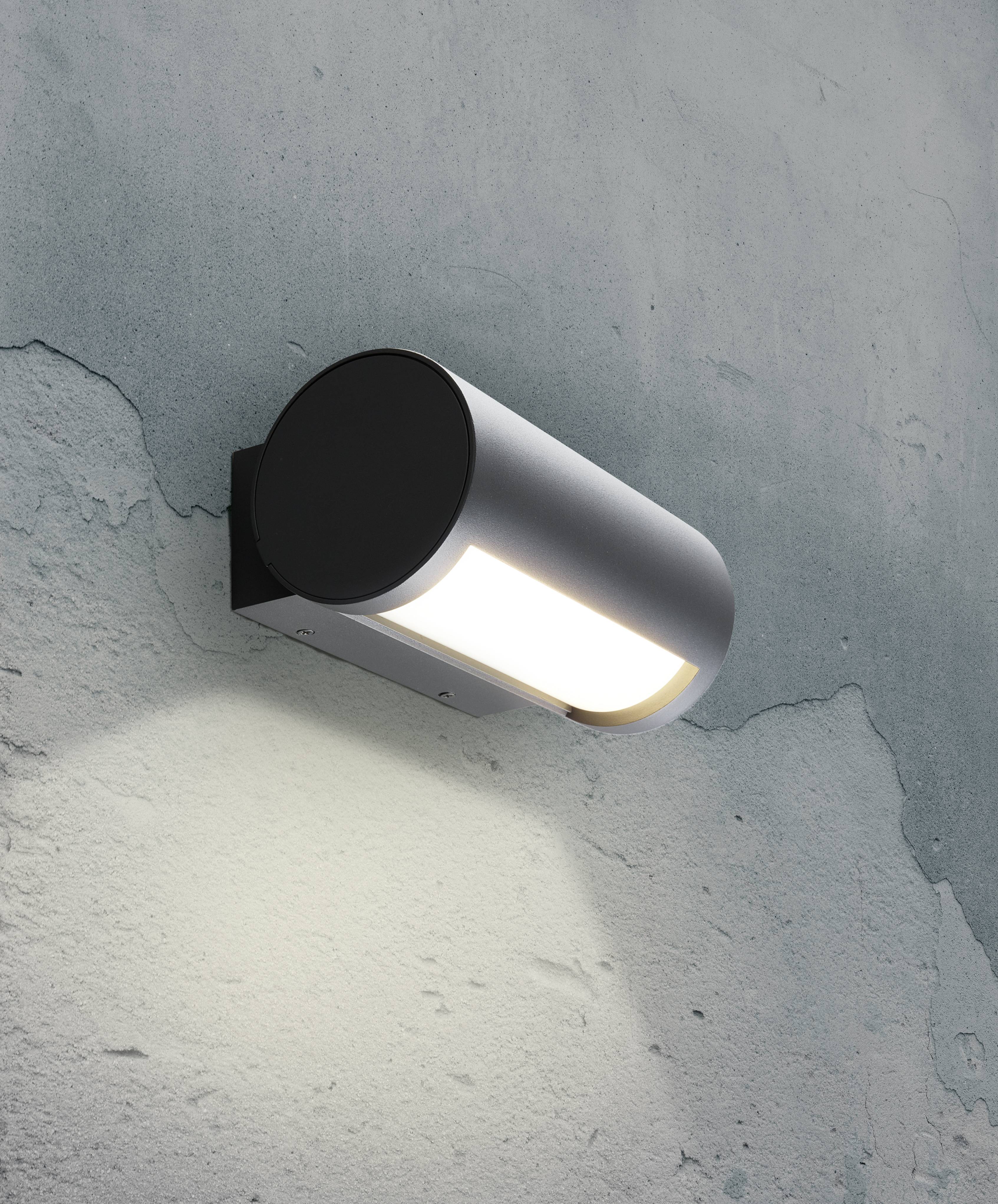 Une lampe murale moderne, cylindrique, avec un support noir, projette une lumière chaleureuse sur une surface murale rugueuse aux tons vert-gris.
