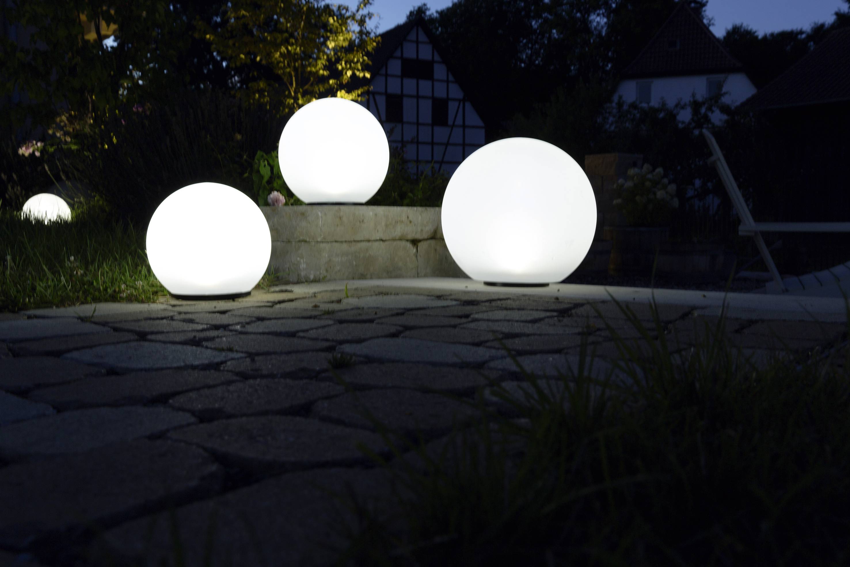 Trois lampes sphériques lumineuses éclairent un chemin de jardin pavé la nuit. En arrière-plan, des arbres et une maison à colombages apparaissent de manière floue.