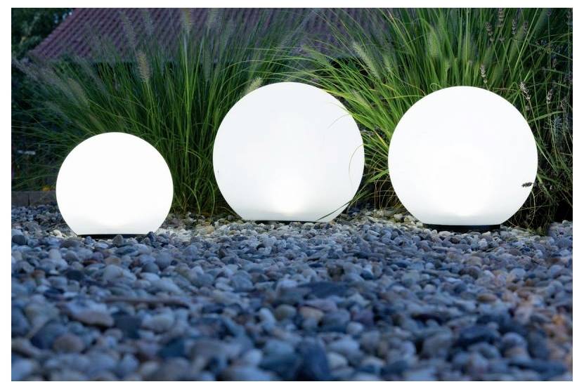 Trois lumières sphériques blanches et brillantes reposent sur du gravier bleu dans un parterre de jardin avec de l'herbe haute derrière elles.