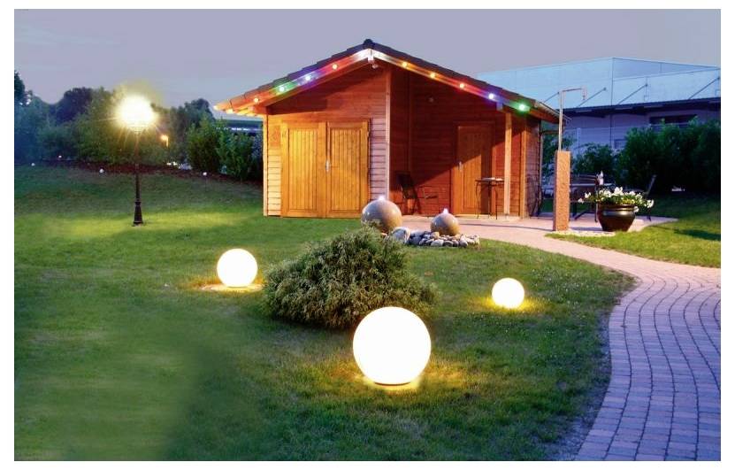 Un abri de jardin en bois avec des luminaires de toit colorés, des lampes sphériques lumineuses sur la pelouse et un chemin de briques courbe au crépuscule.
